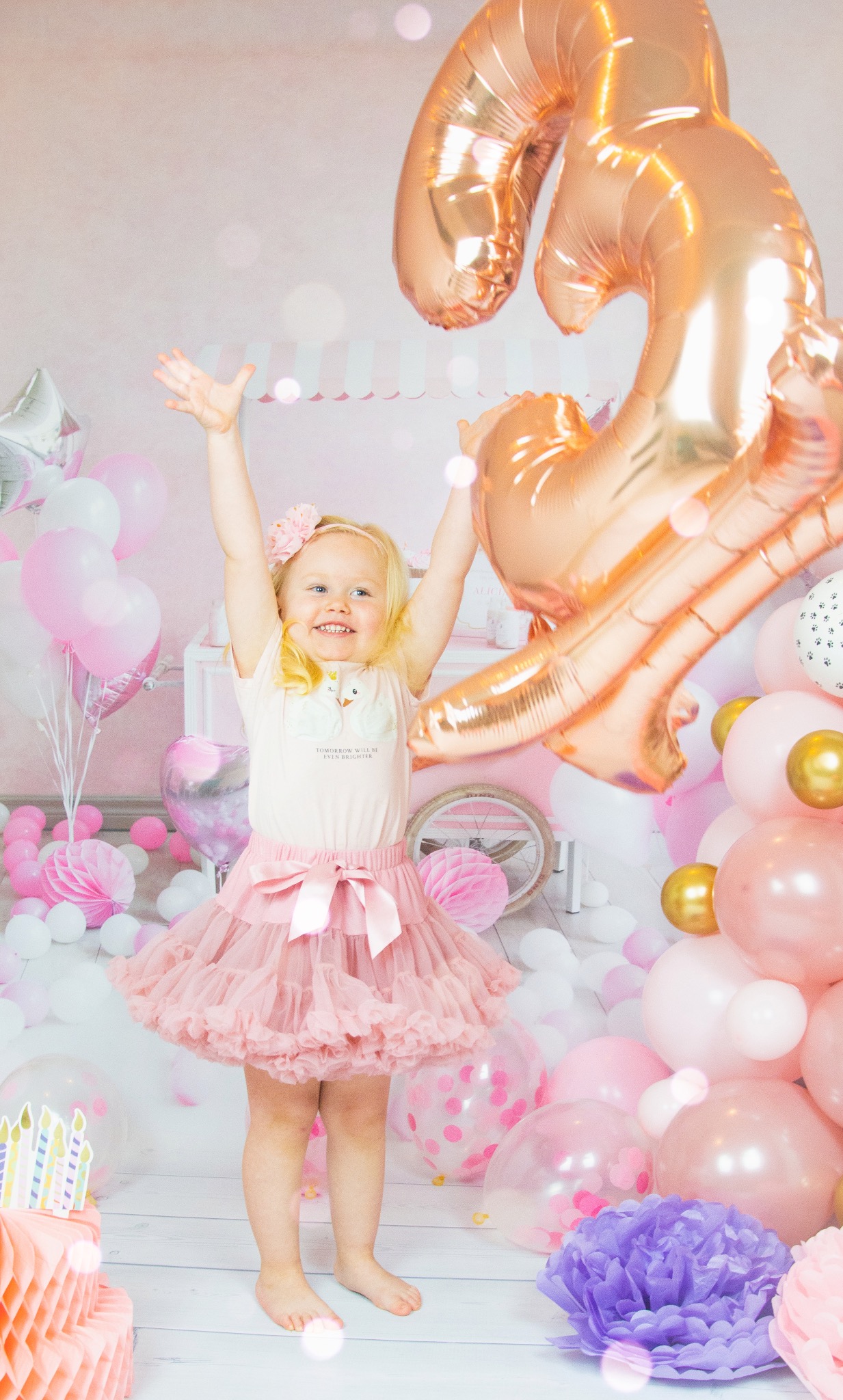 Uśmiechnięta blond dziewczynka w różowej tiulowej spódniczce i bluzce z łabędziami stoi na tle pastelowych balonów i dekoracji urodzinowych, z rękami uniesionymi do góry w geście radości, obok...