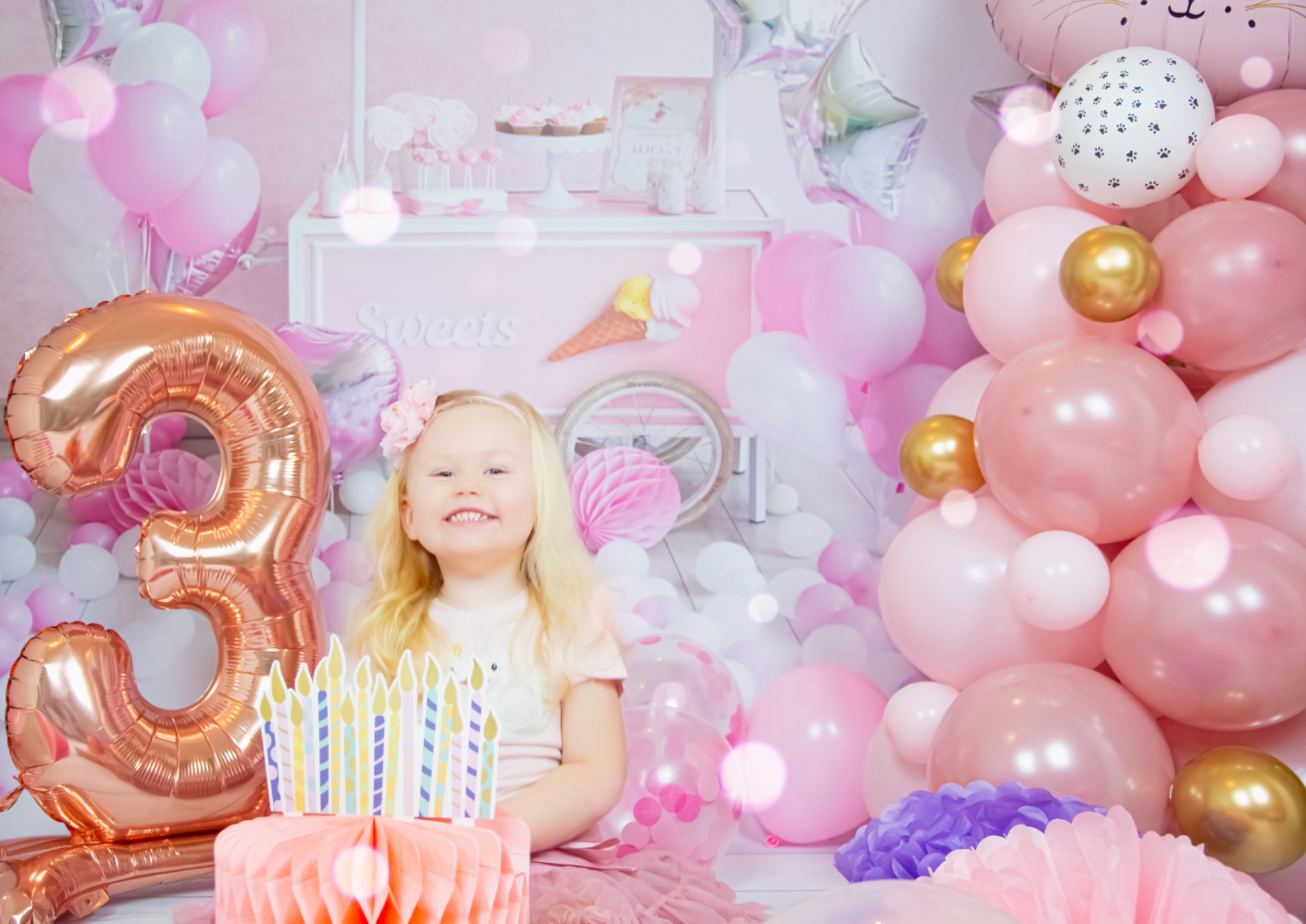 Uśmiechnięta blond dziewczynka w różowej tiulowej spódniczce siedzi na tle dekoracji urodzinowych w odcieniach różu i złota, w tym balon w kształcie cyfry 3, tort z papierowymi świeczkami i wózek...
