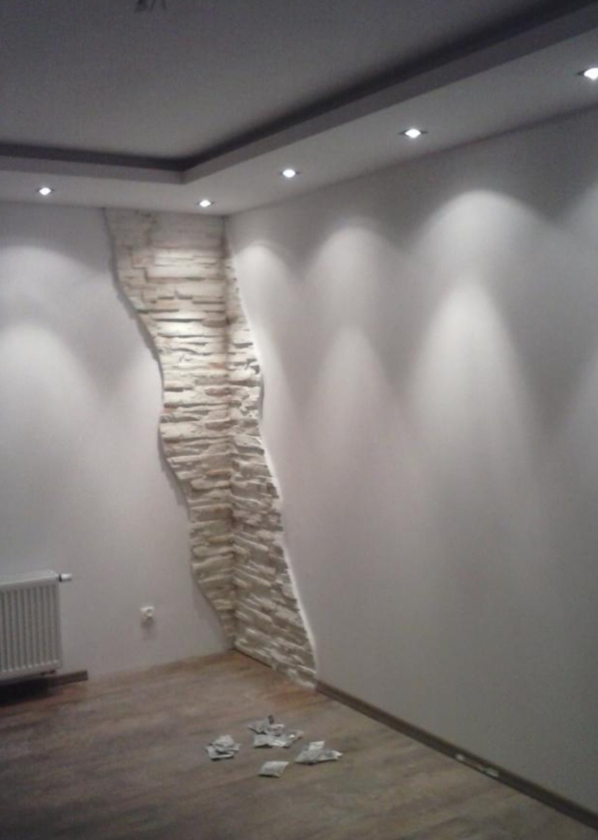 Fragment ściany z dekoracyjną okładziną z kamienia w kształcie nieregularnej kolumny, oświetlona punktowymi lampami sufitowymi, na podłodze leżą resztki materiałów budowlanych, widoczny grzejnik.