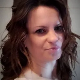 Portret kobiety z brązowymi, falowanymi włosami, delikatnym makijażem i srebrnym naszyjnikiem na tle jasnej ściany.