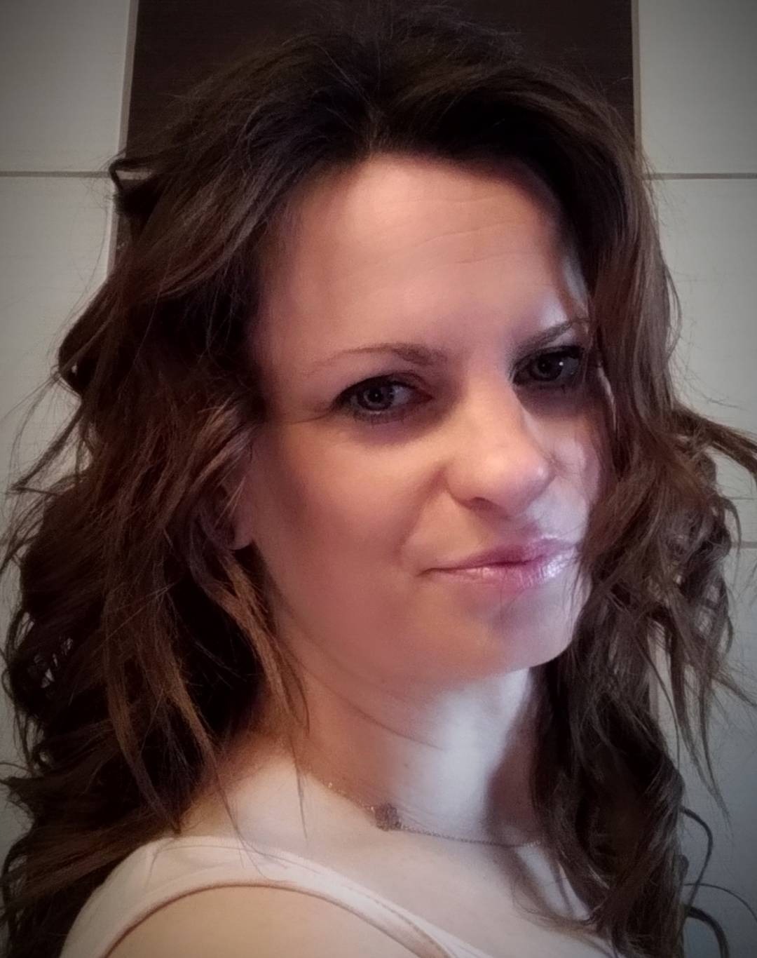 Portret kobiety z brązowymi, falowanymi włosami, delikatnym makijażem i srebrnym naszyjnikiem na tle jasnej ściany.