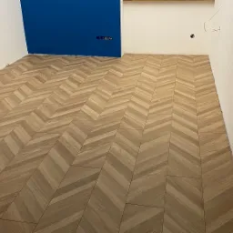 Ułożona w jodełkę jasna podłoga z płytek imitujących drewno w pokoju z pomalowaną na niebiesko ścianą i widocznymi gniazdkami elektrycznymi.