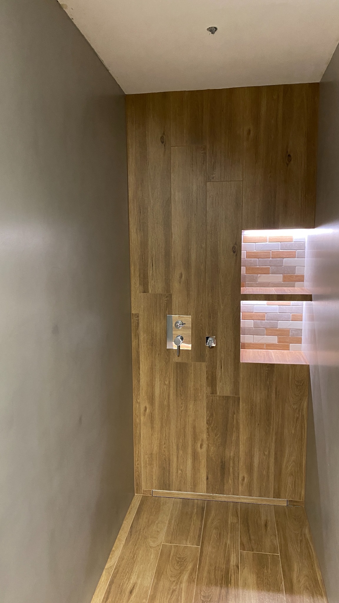 Wykończona łazienka z drewnopodobnymi płytkami na ścianach i podłodze, chromowana bateria podtynkowa i wnęka z mozaiką podświetloną ledami.