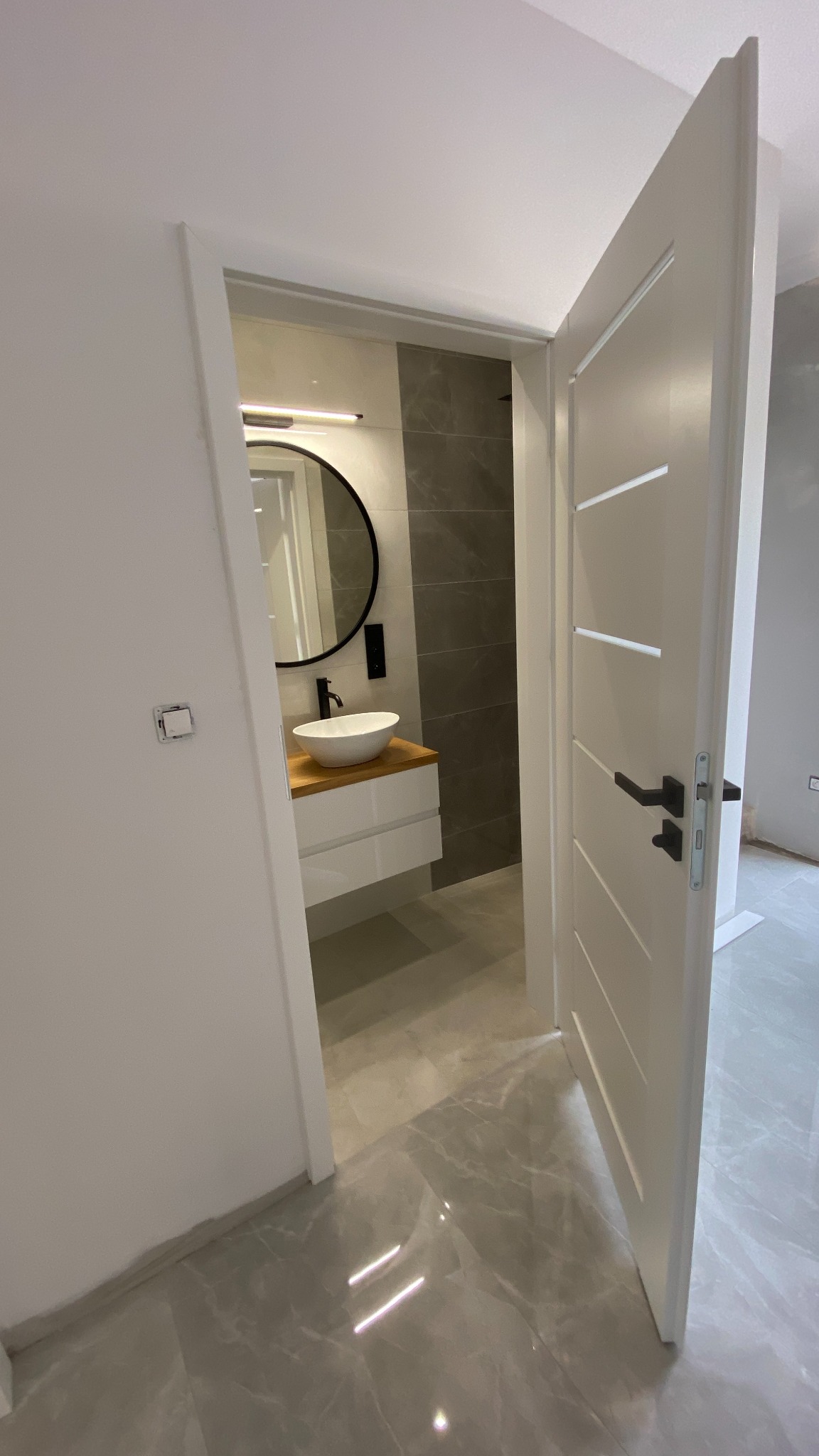 Nowoczesna łazienka z okrągłym lustrem, białą umywalką na drewnianym blacie i szarymi płytkami, widoczna przez otwarte białe drzwi z poziomymi frezami i czarną klamką.