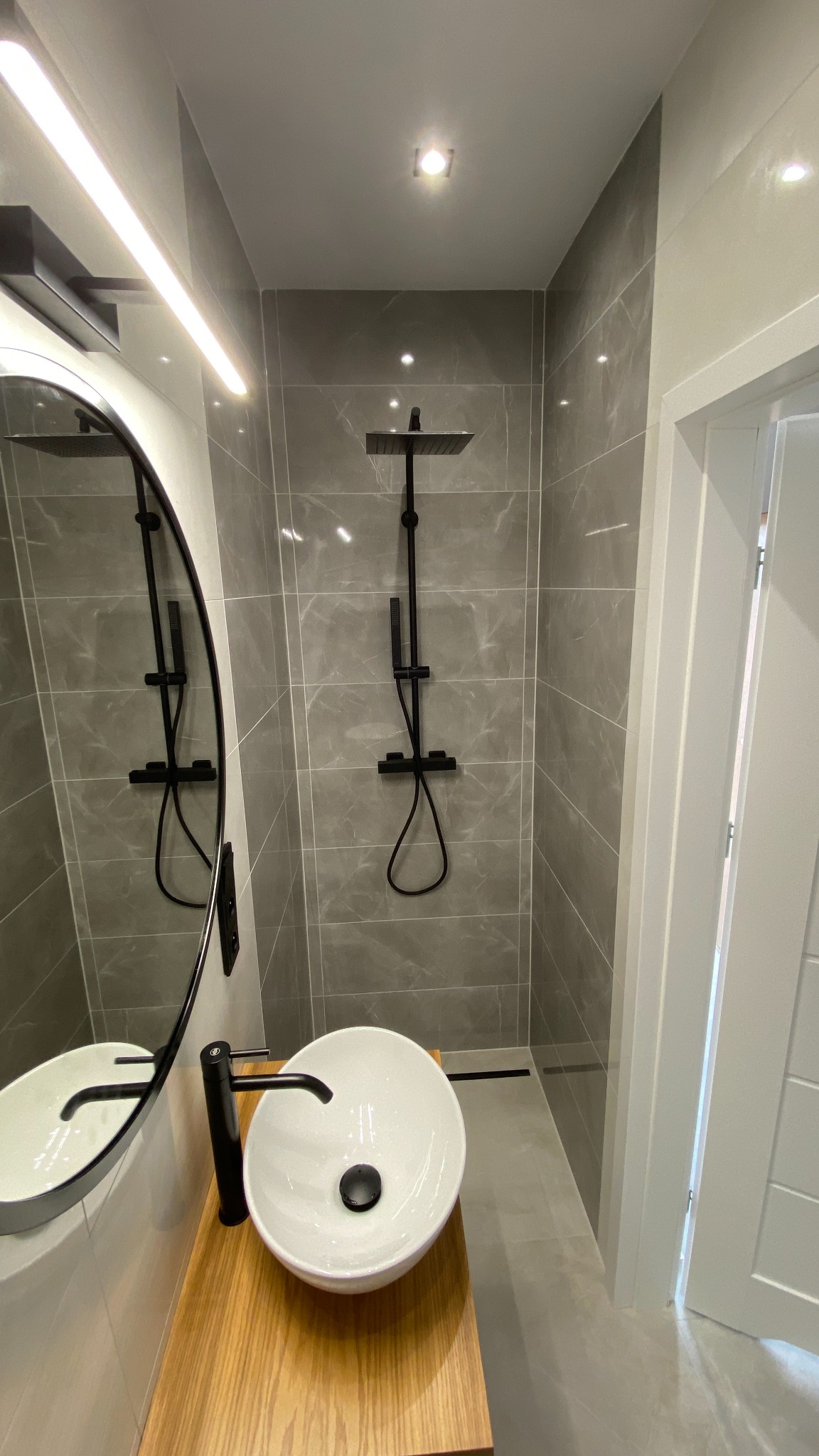 Nowoczesna łazienka z szarymi płytkami imitującymi marmur, czarną armaturą prysznicową i okrągłym lustrem z podświetleniem LED nad umywalką nablatową na drewnianym blacie.