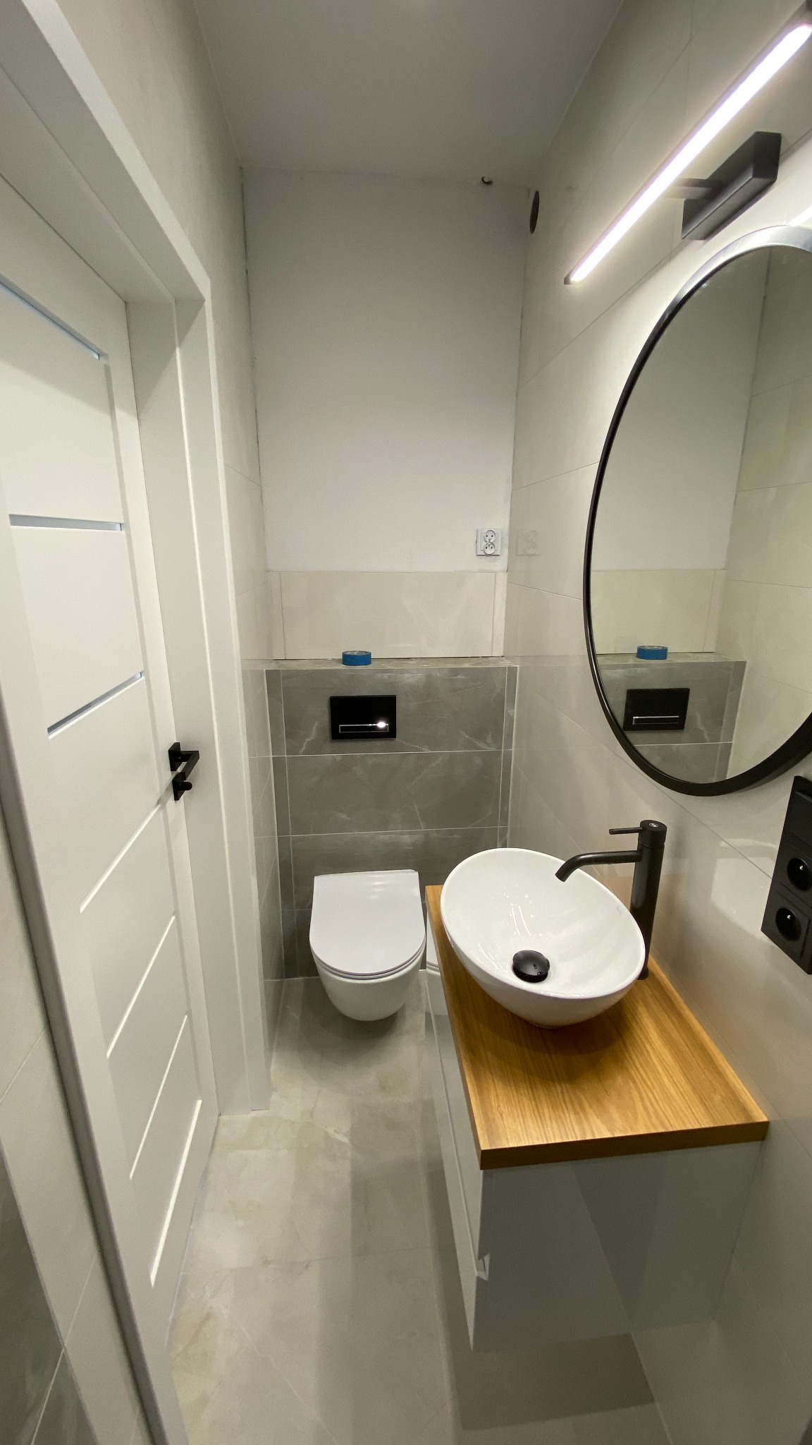 Nowoczesna, mała łazienka z wiszącą toaletą, owalną umywalką nablatową na drewnianym blacie i okrągłym lustrem z czarną ramą, oświetlona listwą LED.