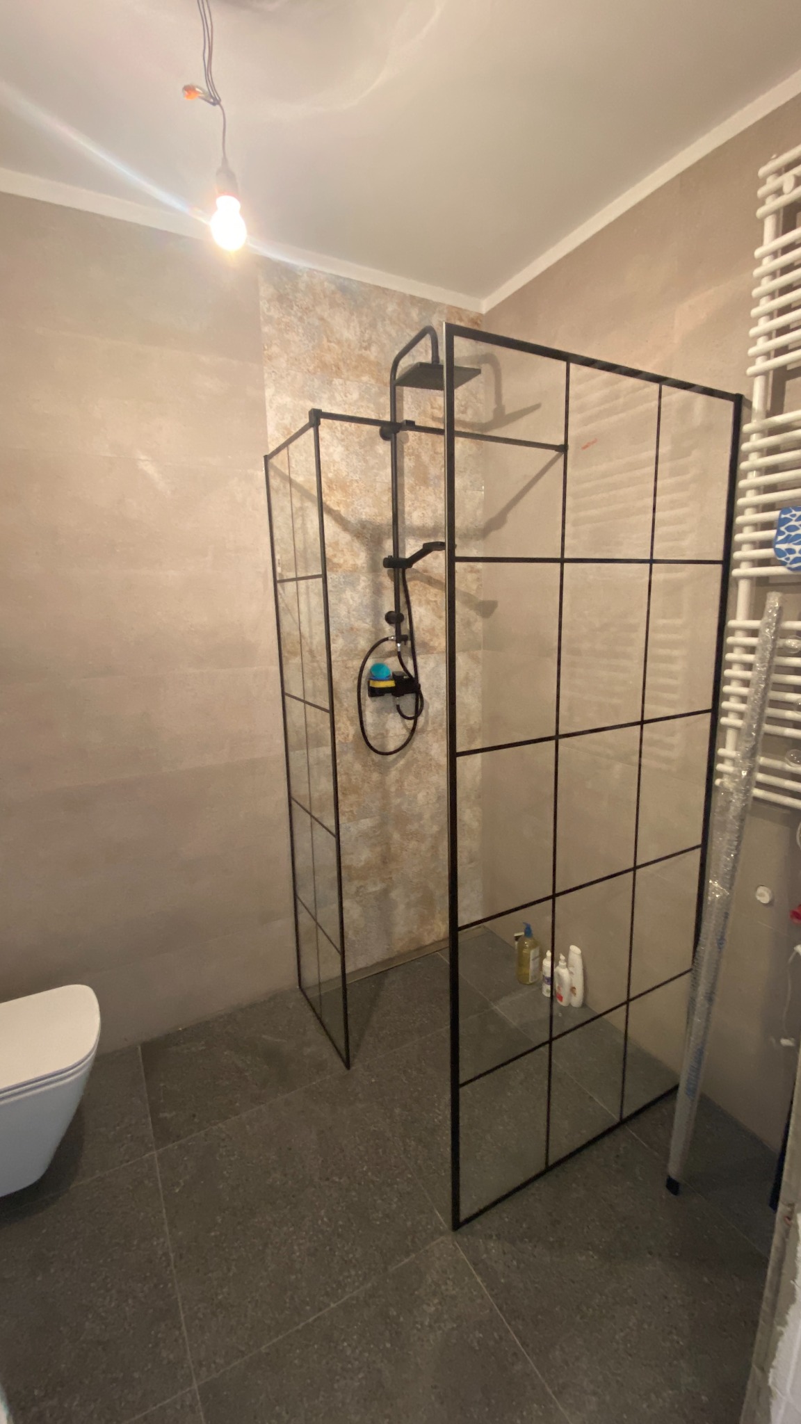 Nowoczesna kabina prysznicowa z czarnymi profilami i szklanymi ściankami, widoczny biały grzejnik drabinkowy i toaleta w tle, szare płytki podłogowe, surowy styl wykończenia.