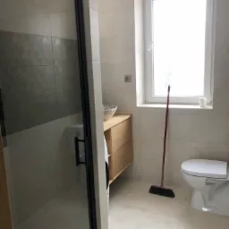 Nowoczesna łazienka z kabiną prysznicową z czarnymi profilami, umywalką nablatową na drewnianej szafce, toaletą i czerwoną miotłą opartą o ścianę pod oknem.