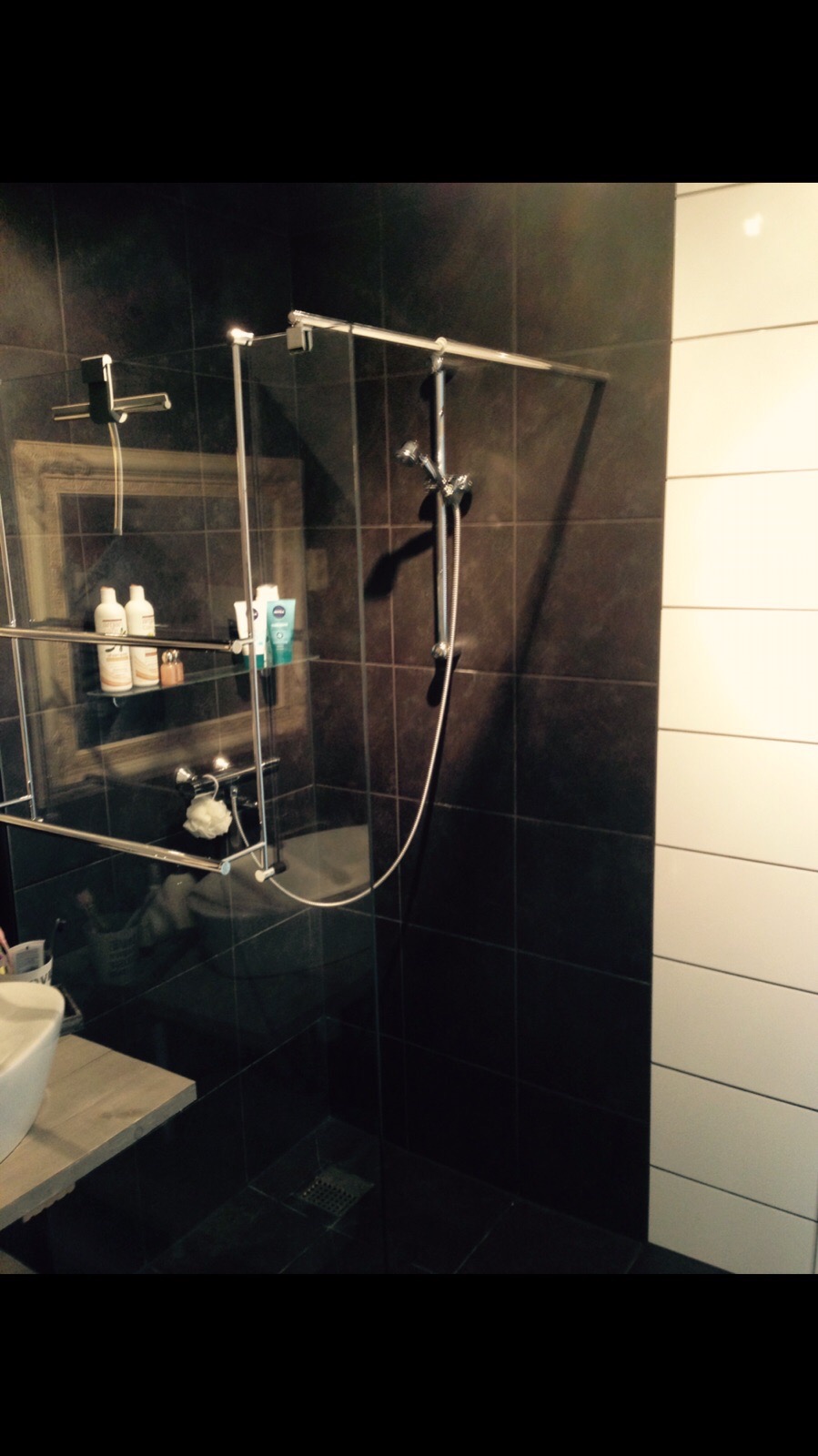 Nowoczesna kabina prysznicowa z brązowymi płytkami i chromowaną armaturą, półki na kosmetyki, umywalka z drewnianym blatem po lewej stronie.