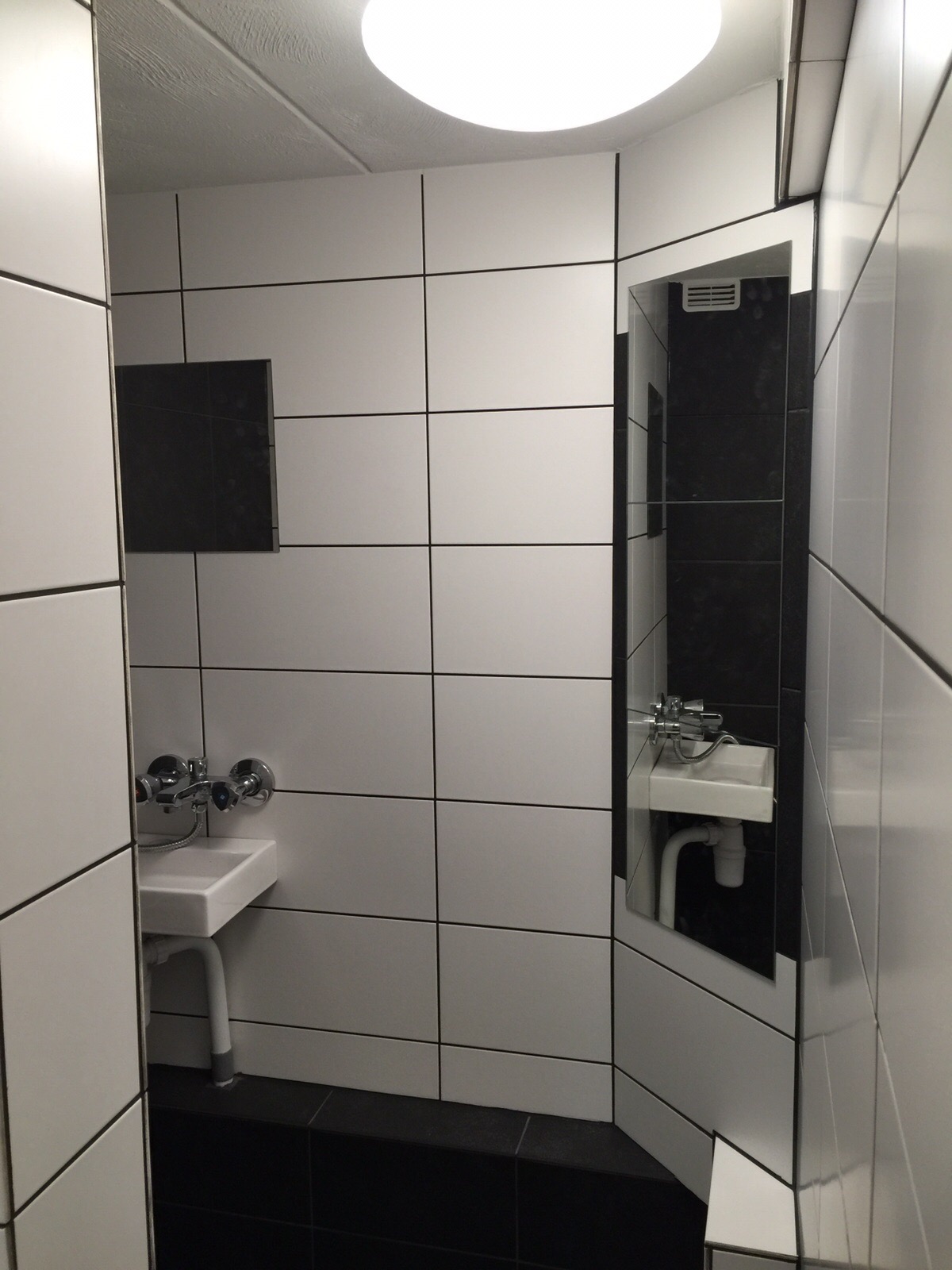 Mała, wykafelkowana łazienka z biało-czarnymi płytkami, małą białą umywalką i lustrem z widocznym odbiciem umywalki.