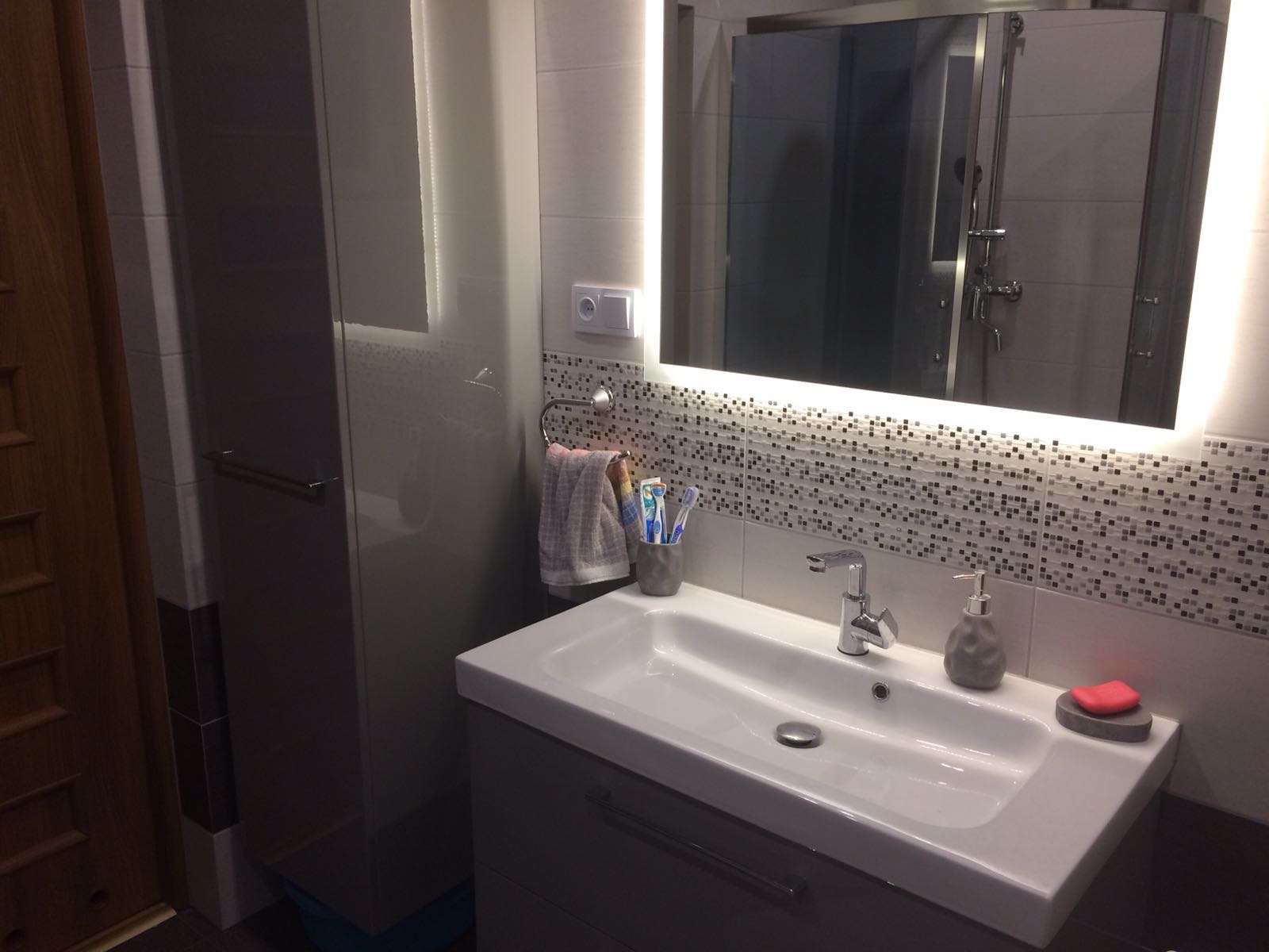 Nowoczesna łazienka z podświetlanym lustrem, umywalką nablatową, mozaiką na ścianie i szarą szafką pod umywalką.