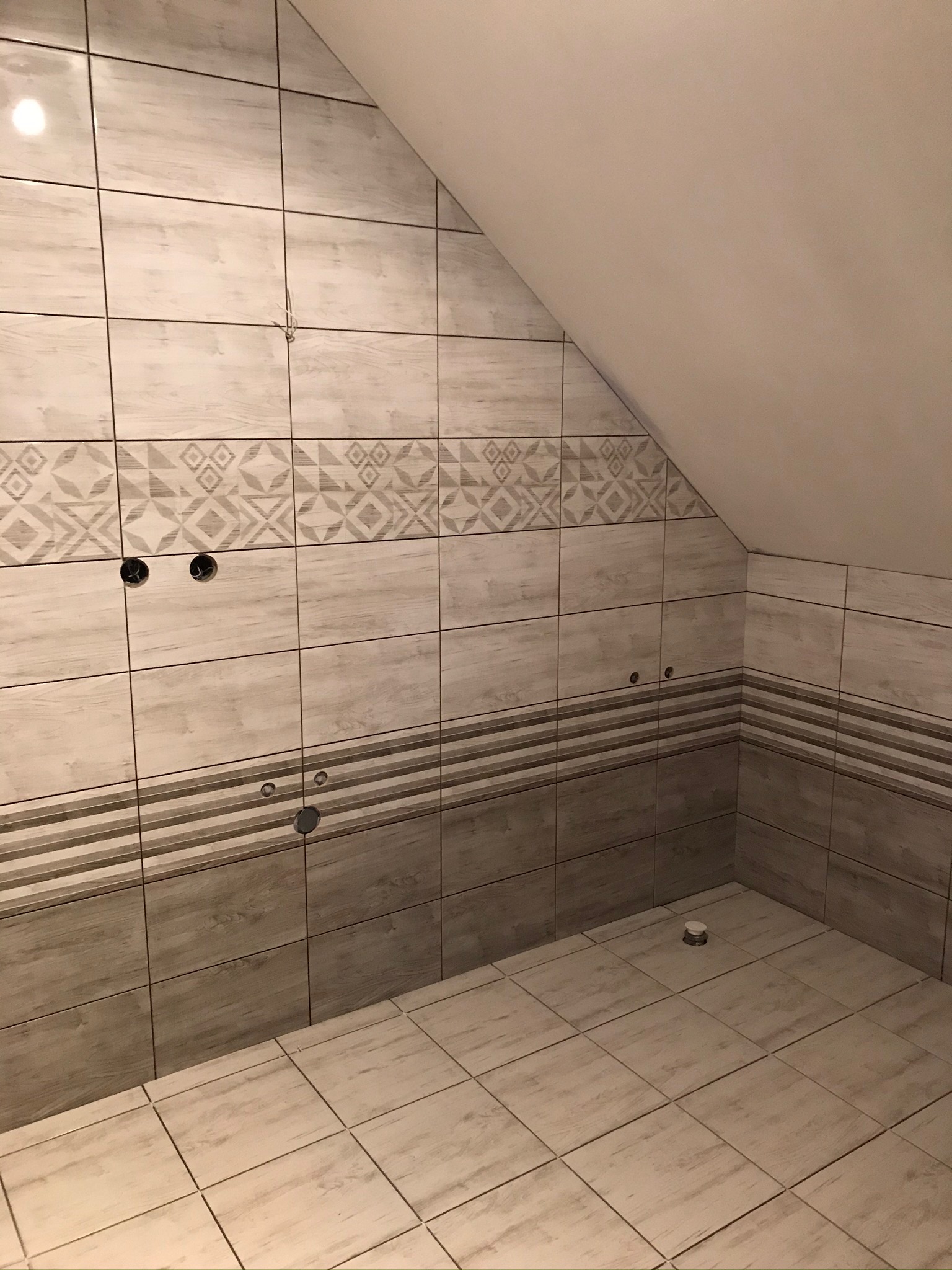 Wykończona łazienka na poddaszu ze skośnym sufitem, wyłożona płytkami w odcieniach beżu i szarości z dekoracyjnymi paskami i wzorami, widoczne otwory na armaturę.