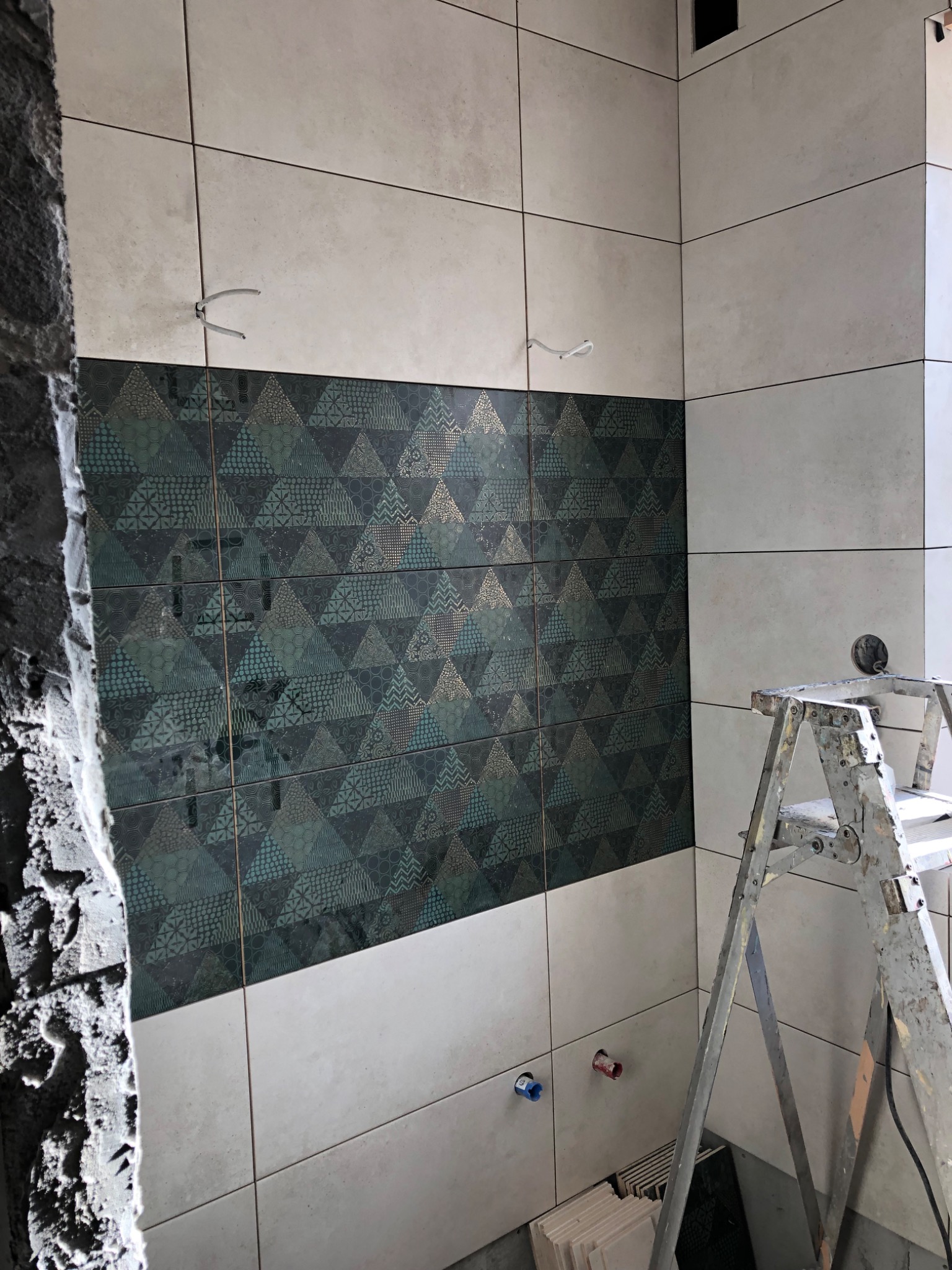 Układanie płytek w łazience: fragment ściany z dekoracyjnymi, zielono-złotymi płytkami o wzorze geometrycznym, sąsiadującymi z jasnymi, beżowymi płytkami. Widoczne wystające kable, rury...