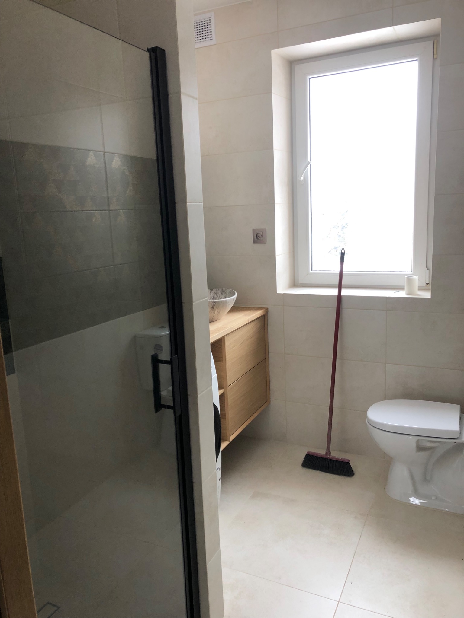 Nowoczesna łazienka z kabiną prysznicową z czarnymi profilami, umywalką nablatową na drewnianej szafce, toaletą i czerwoną miotłą opartą o ścianę pod oknem.