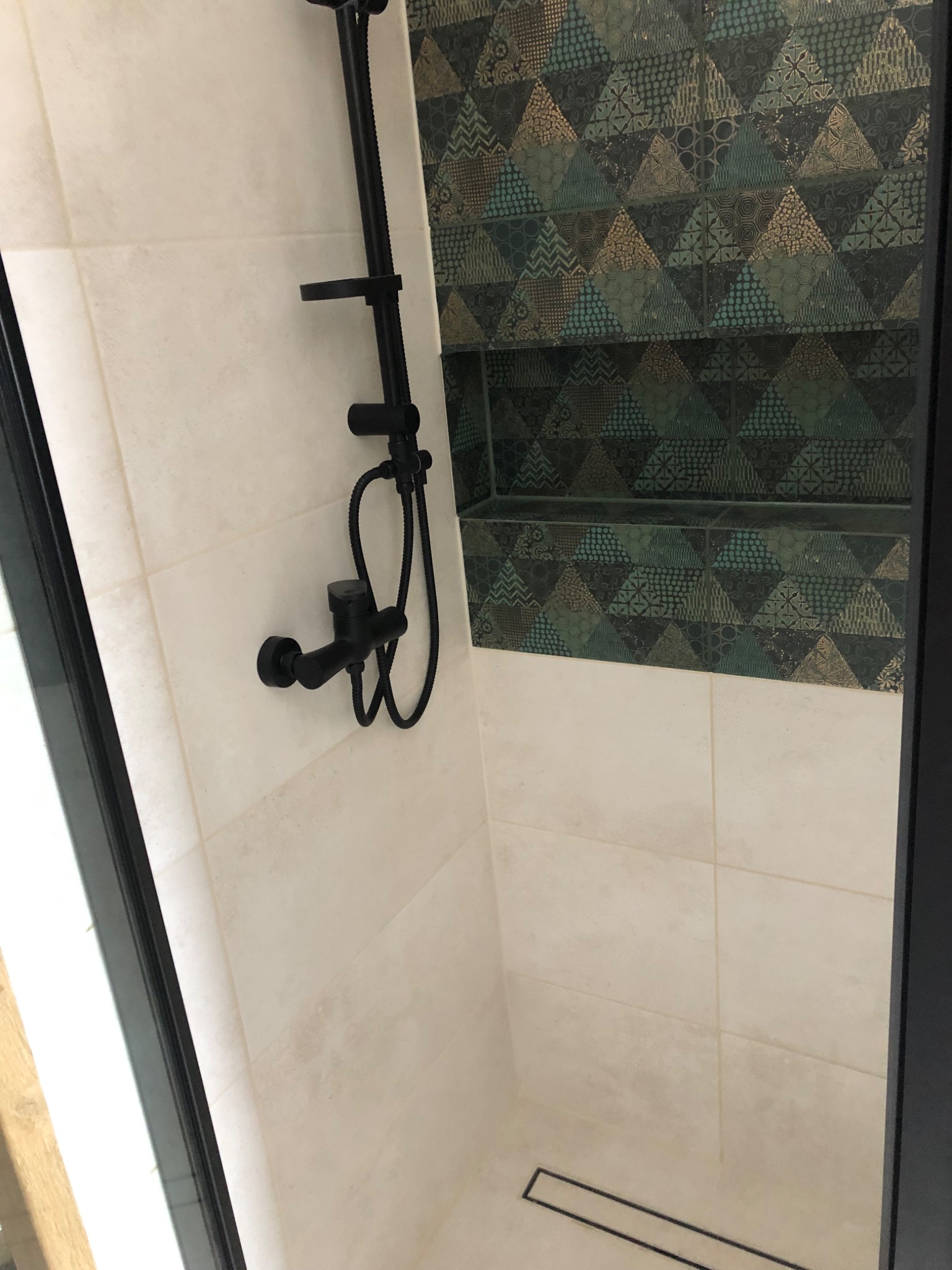 Wykończona kabina prysznicowa z czarną armaturą i geometrycznymi płytkami w odcieniach zieleni i złota we wnęce, jasne płytki na pozostałych ścianach.