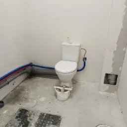 Biała toaleta kompaktowa z podłączonymi rurami wodnymi i odpływowymi w trakcie montażu w surowym, wykończonym na biało pomieszczeniu. Widoczne narzędzia w wiadrze, rury w ścianie i otwór w ścianie...