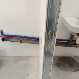 Wnętrze łazienki w trakcie remontu: widoczne rury instalacji wodnej (czerwone i niebieskie) oraz kanalizacyjnej (szare) wyprowadzone ze ściany, obok umywalka i metalowy profil ściany działowej,...