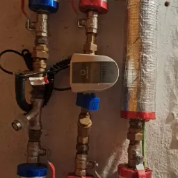 Pionowe rury instalacyjne z izolacją aluminiową, zawory z czerwonymi i niebieskimi uchwytami, oraz elektroniczny sterownik zamontowany na jednej z rur.