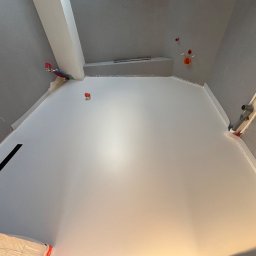 Sicher Bautechnik - Świeżo wylana, jasnoszara podłoga w pomieszczeniu w trakcie remontu, widoczne wystające rury i instalacje na ścianach.