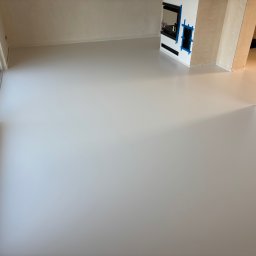 Sicher Bautechnik - Jasnoszara, gładka podłoga w nowym pomieszczeniu z dużym oknem i przygotowanym miejscem na kominek, zabezpieczonym niebieską taśmą malarską.