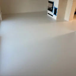 Jasnoszara, gładka podłoga w nowym pomieszczeniu z dużym oknem i przygotowanym miejscem na kominek, zabezpieczonym niebieską taśmą malarską.