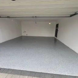 Gładka, szara posadzka w garażu z widocznymi drobinkami, świeżo po aplikacji. Białe ściany, betonowy sufit i fragment bramy garażowej. Widoczne kable i instalacje.