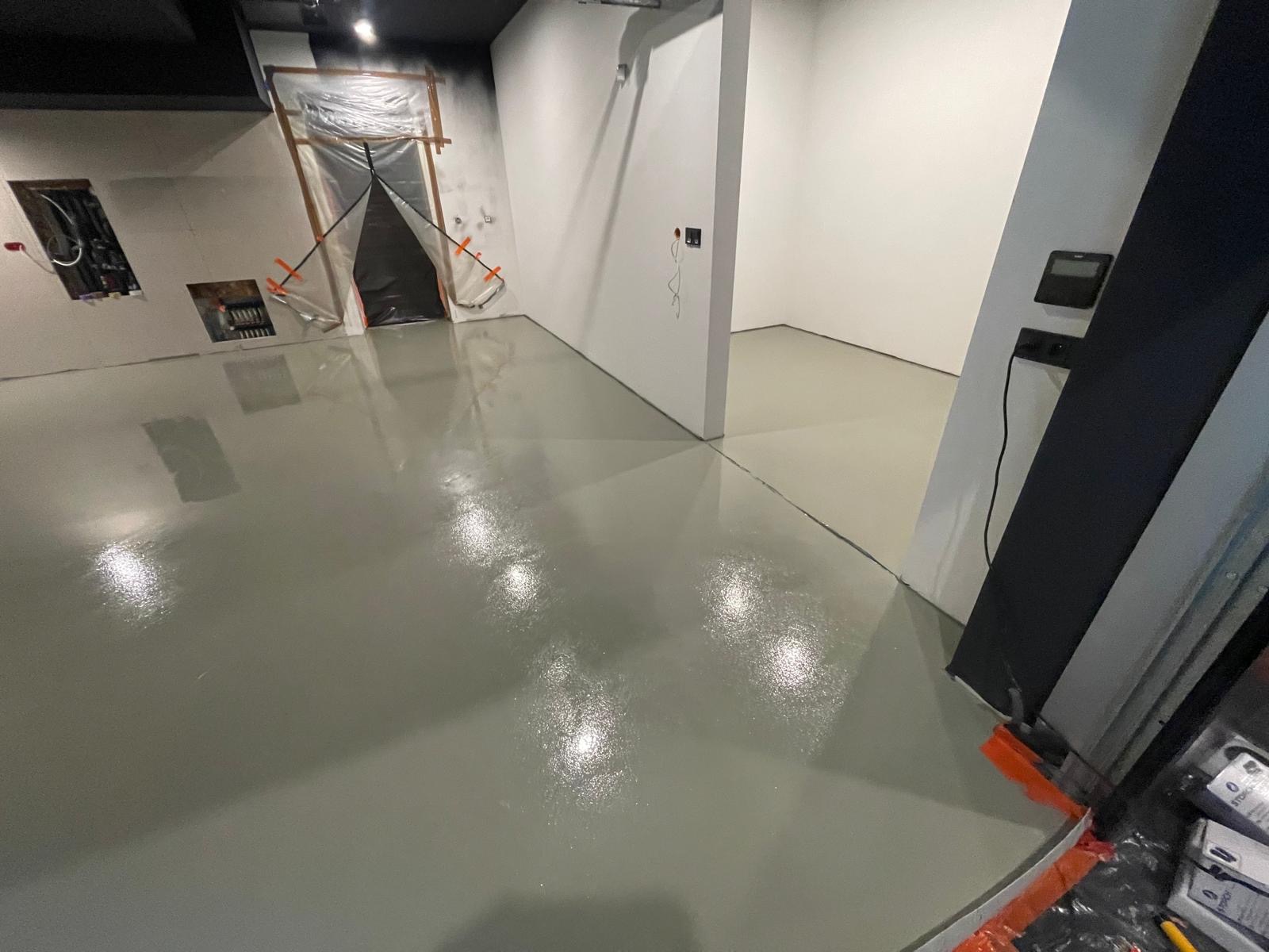 Świeżo wylana, jasnoszara posadzka żywiczna w pomieszczeniu z przygotowanymi otworami w ścianie i zabezpieczonym wejściem folią malarską.