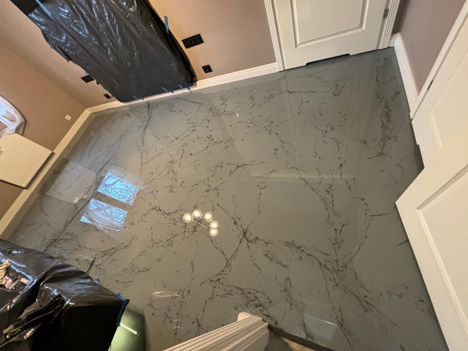 Szara, błyszcząca posadzka z efektem marmuru, zabezpieczona folią malarską w części pomieszczenia, widoczne odbicie okna i lampy w powierzchni.
