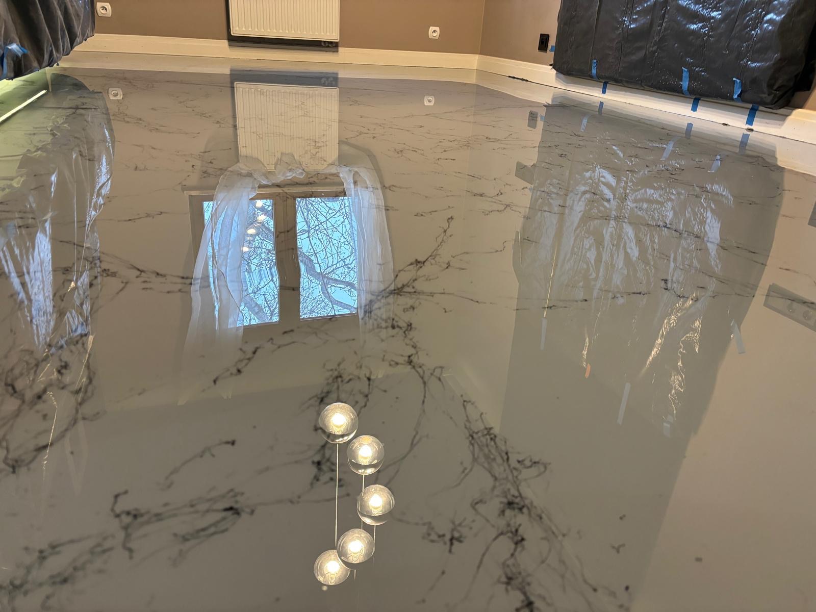 Błyszcząca, szara podłoga z efektem marmuru, odbijająca okno z białymi firankami i wiszącą lampę z pięcioma kulami, zabezpieczona folią malarską przy ścianach.