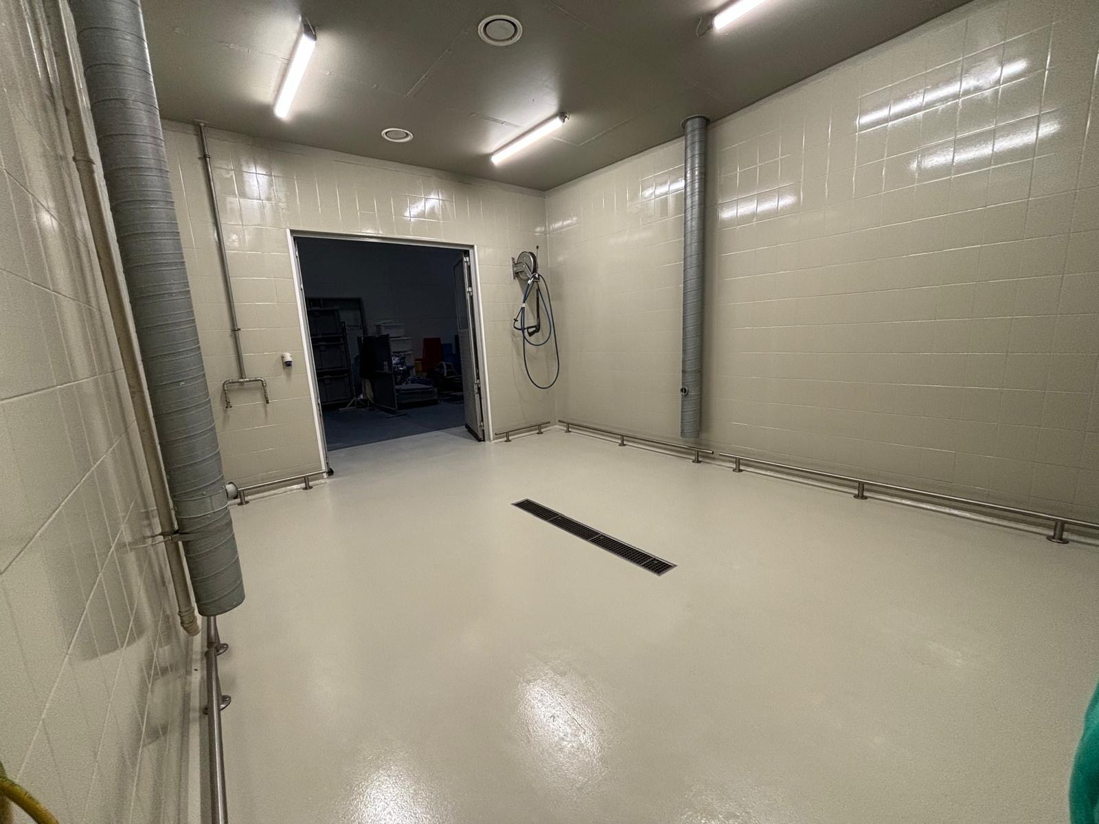 Jasna, gładka posadzka w pomieszczeniu z wyłożonymi kafelkami ścianami, widoczny odpływ liniowy i rury wentylacyjne.