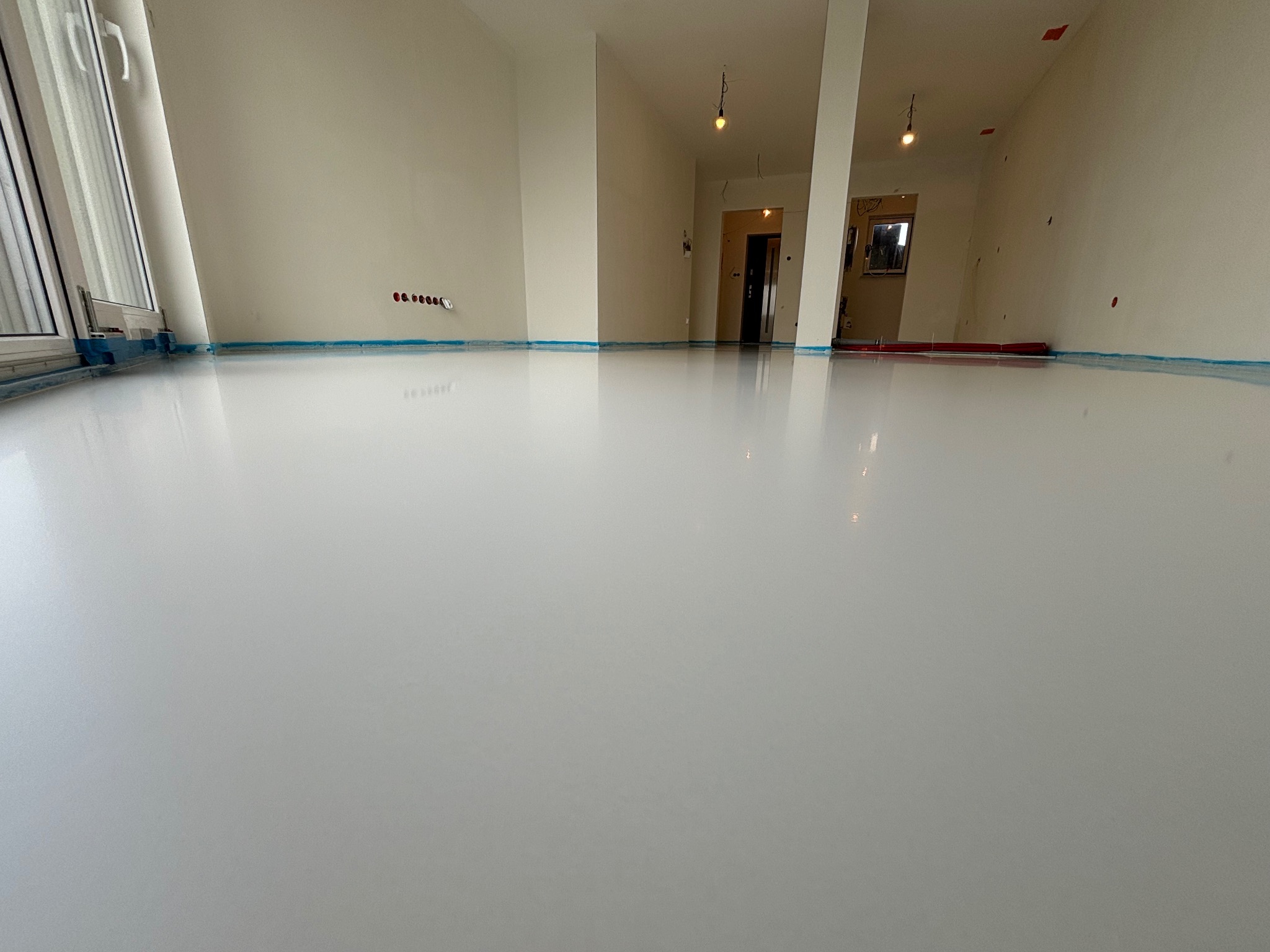 Biała, błyszcząca posadzka żywiczna w jasnym, pustym pomieszczeniu z oknem i widocznymi gniazdkami elektrycznymi na ścianie, zabezpieczona niebieską taśmą malarską przy krawędziach.
