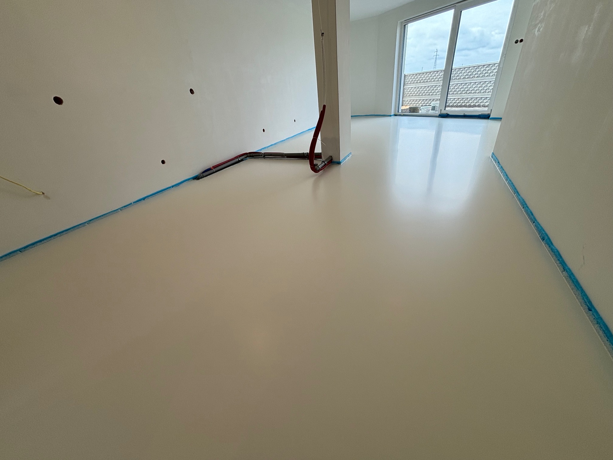 Jasna, błyszcząca podłoga w nowym pomieszczeniu, zabezpieczona niebieską taśmą malarską przy ścianach, widoczne rurki instalacji grzewczej na podłodze i otwory w ścianie.