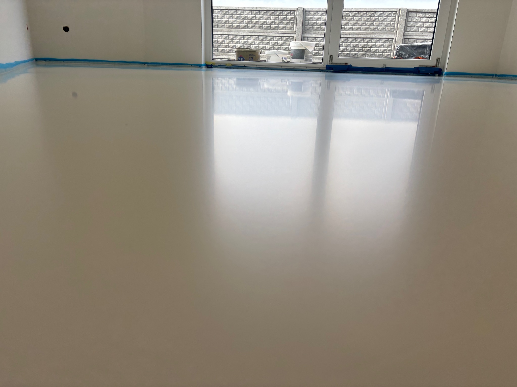 Beżowa, błyszcząca posadzka w jasnym pomieszczeniu z dużym oknem, zabezpieczona niebieską taśmą malarską przy ścianach, z widocznymi pojemnikami na parapecie.