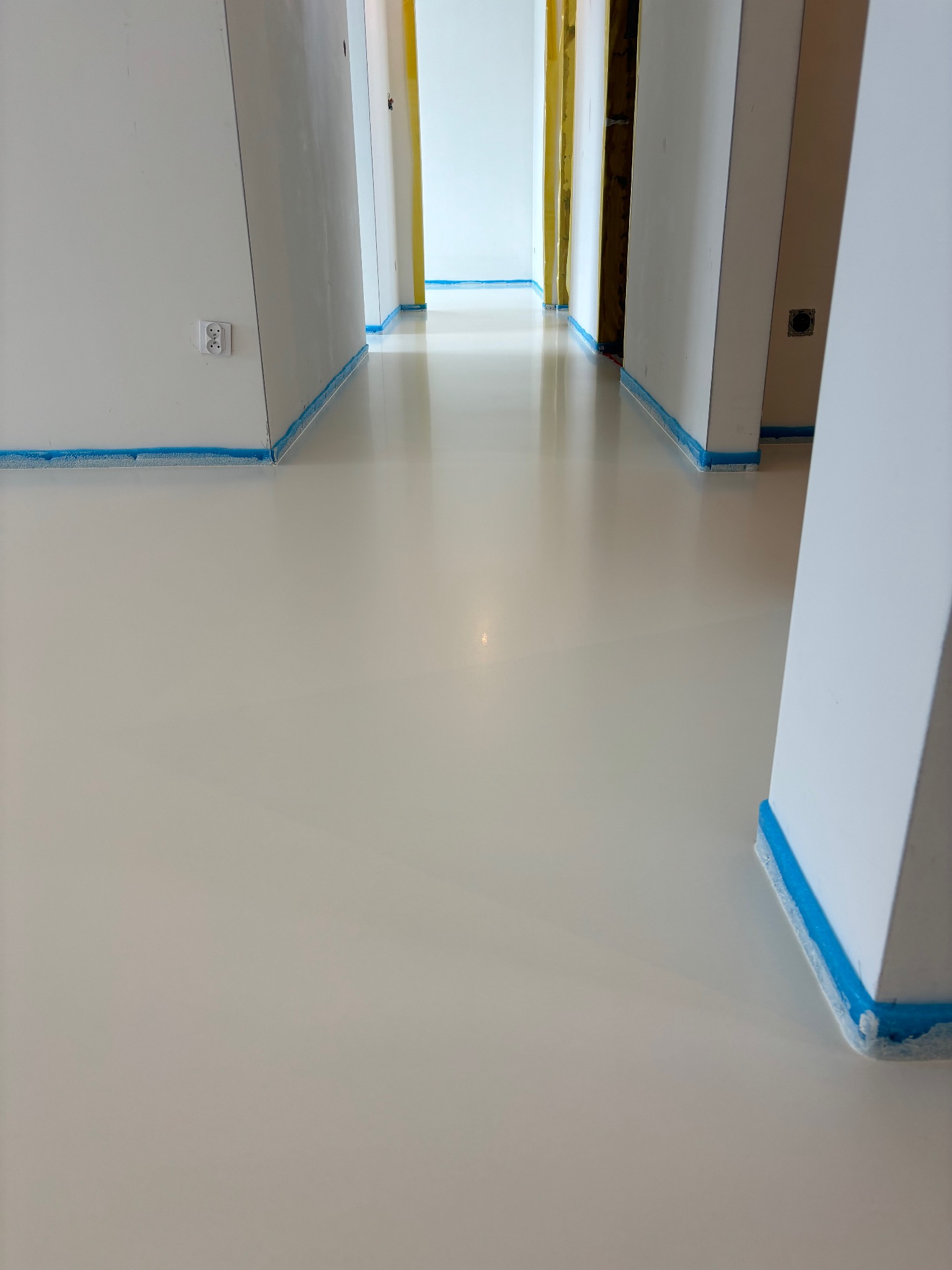 Biała, lśniąca posadzka w jasnym korytarzu, zabezpieczona niebieską taśmą malarską przy ścianach i ościeżnicach.