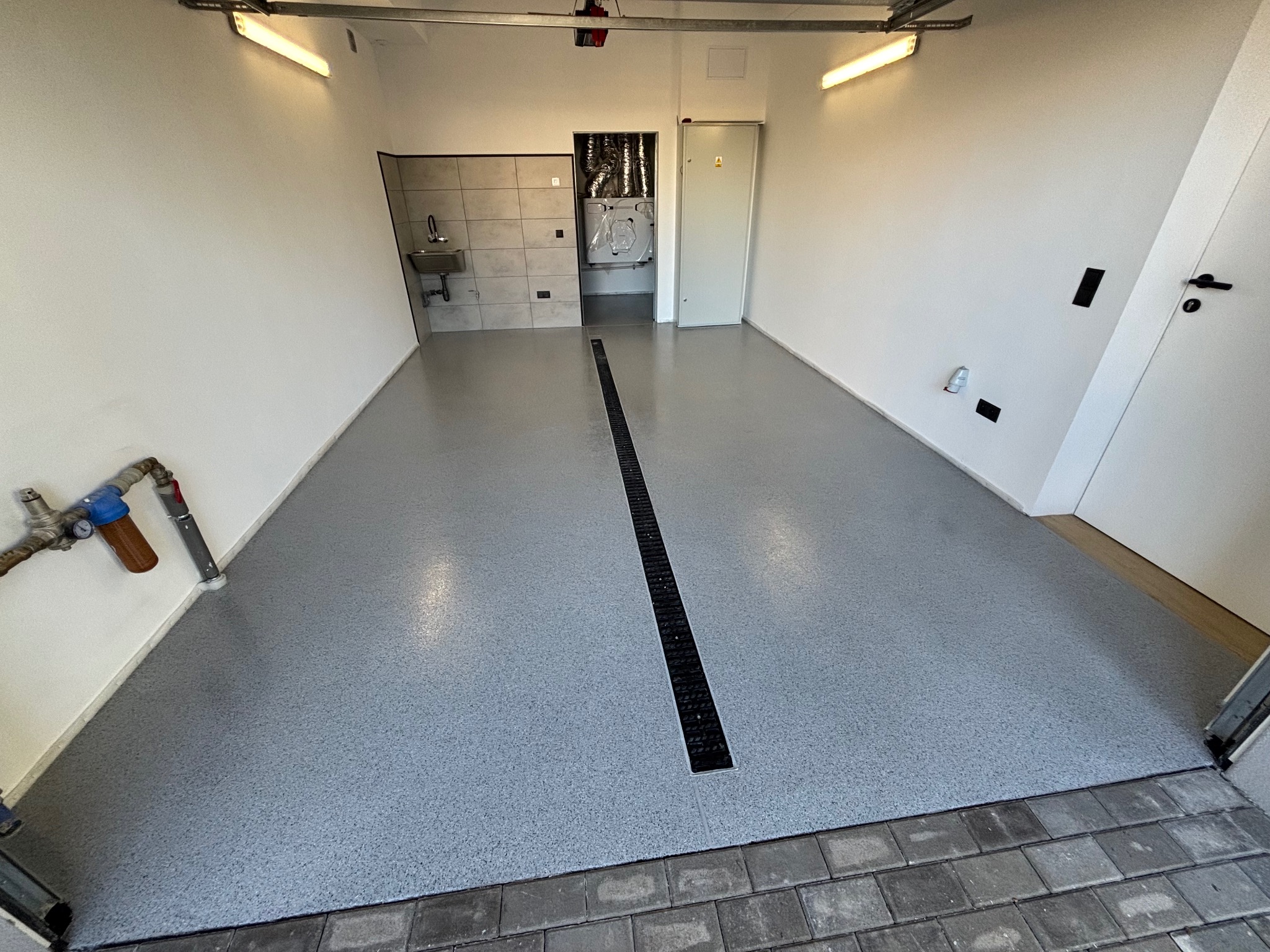 Jasnoszara posadzka w garażu z odpływem liniowym pośrodku, widoczne instalacje wodne przy ścianie, białe ściany i oświetlenie sufitowe.