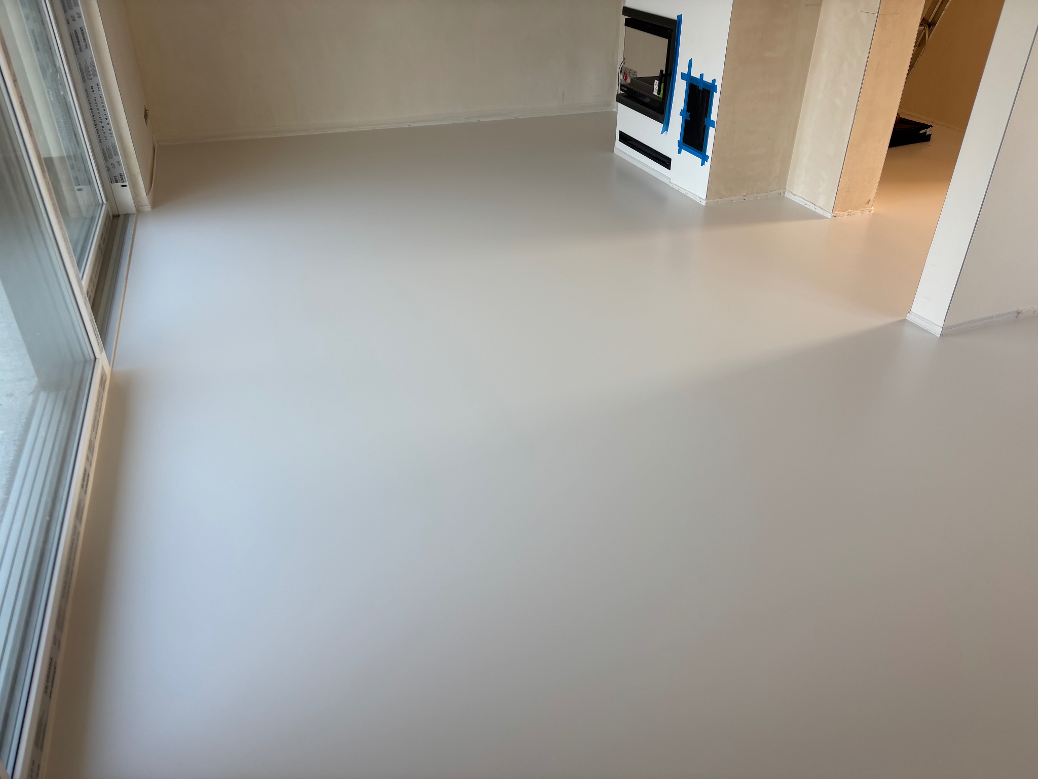 Jasnoszara, gładka podłoga w nowym pomieszczeniu z dużym oknem i przygotowanym miejscem na kominek, zabezpieczonym niebieską taśmą malarską.