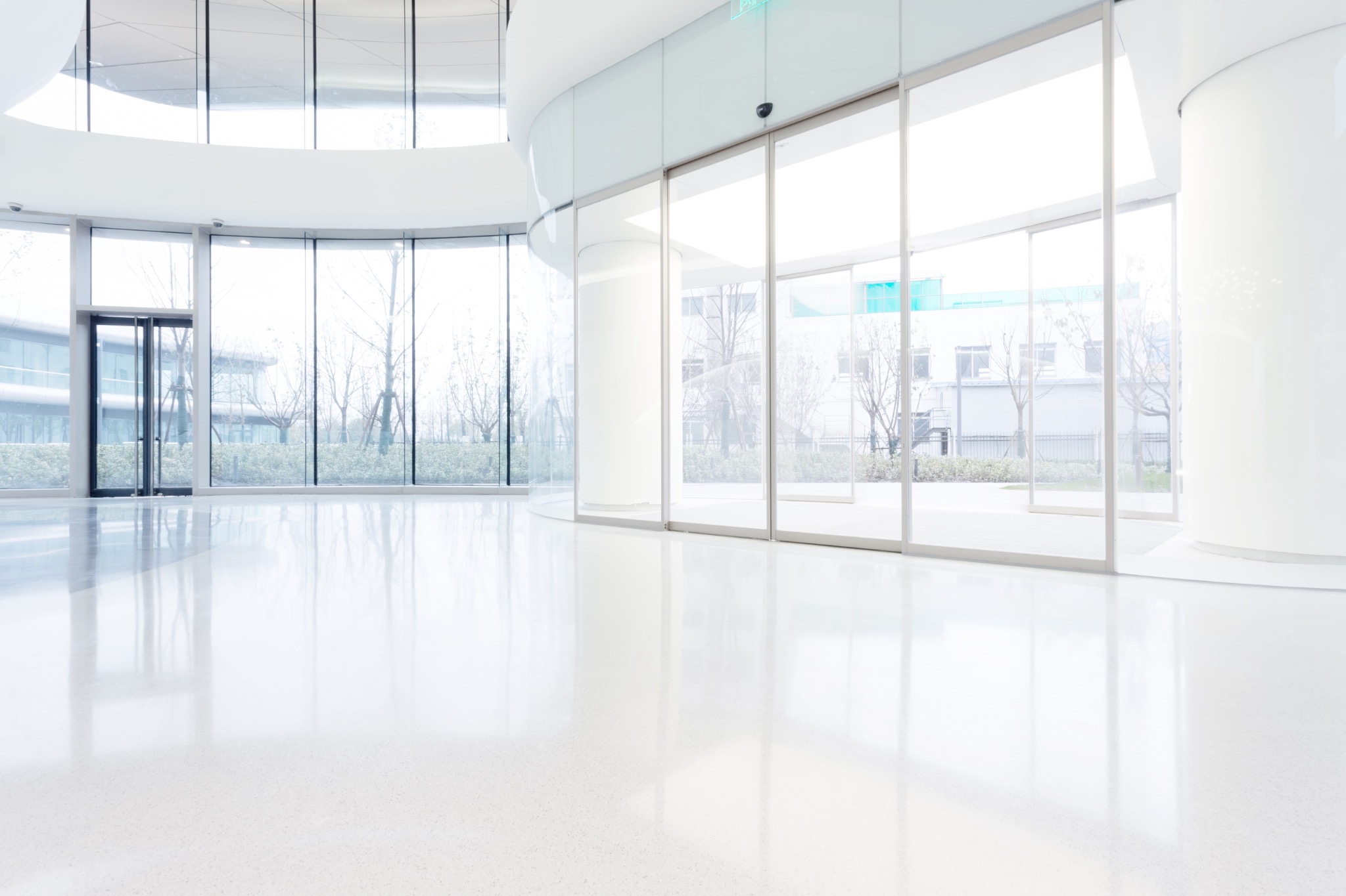 Białe, przestronne wnętrze z błyszczącą posadzką i panoramicznymi oknami wychodzącymi na zieleń, widoczne odbicia w podłodze potęgują wrażenie czystości.