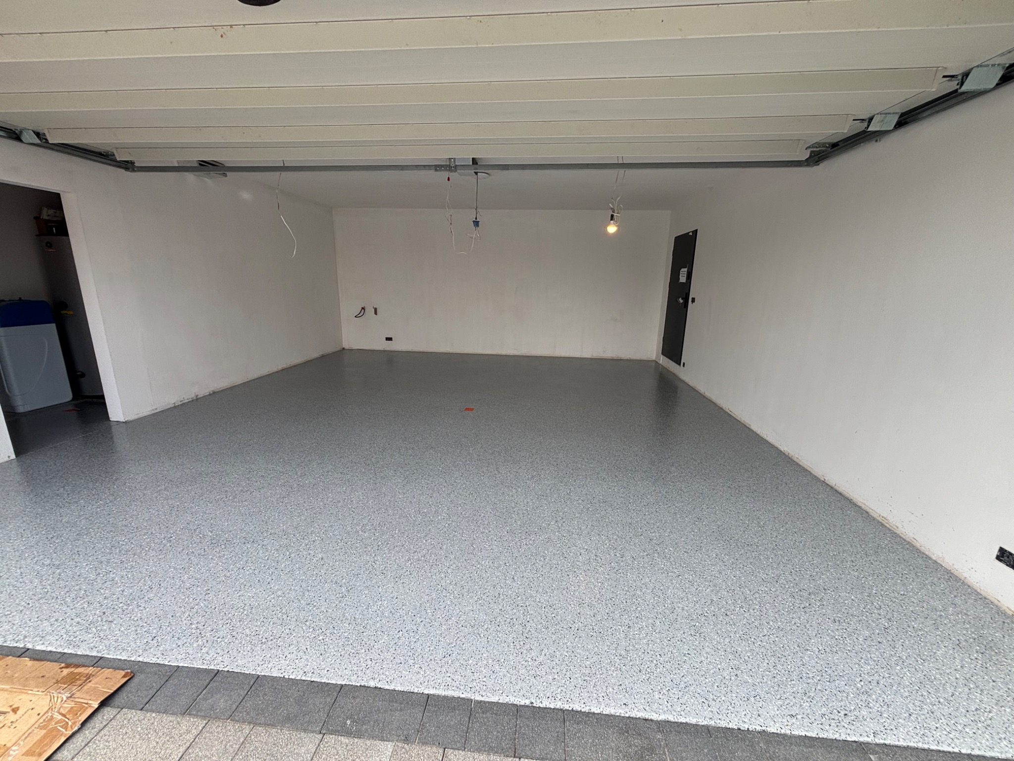 Gładka, szara posadzka w garażu z widocznymi drobinkami, świeżo po aplikacji. Białe ściany, betonowy sufit i fragment bramy garażowej. Widoczne kable i instalacje.