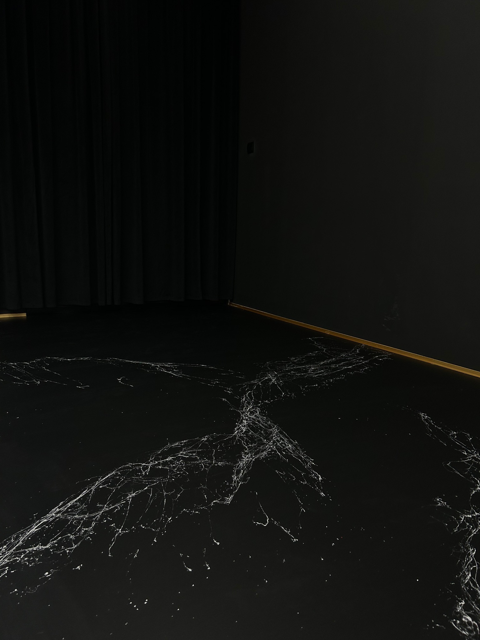 Czarne wykończenie podłogi z białymi żyłkami, imitujące marmur, przy czarnej ścianie i zasłonie. Widoczne złote listwy przypodłogowe.