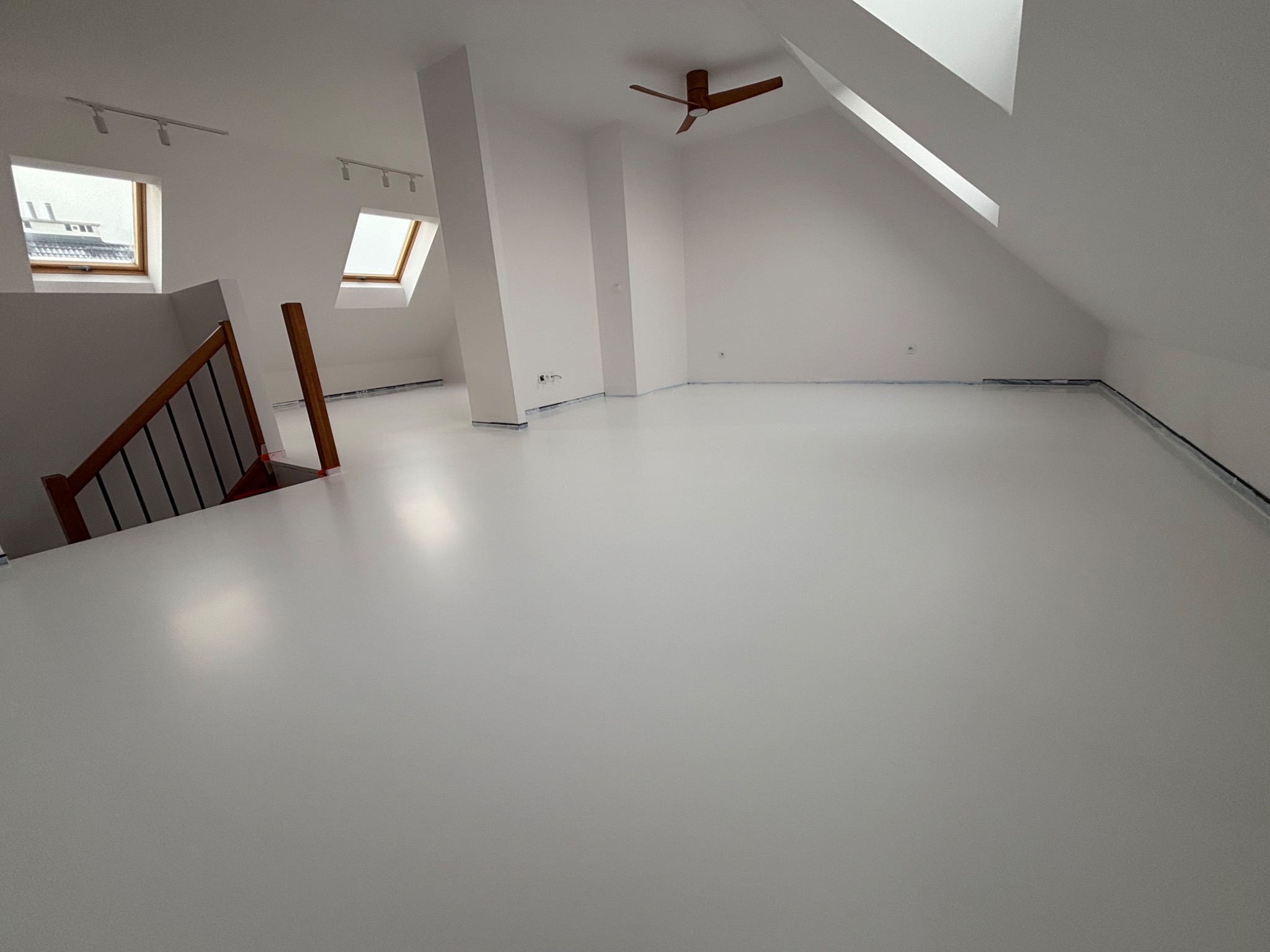 Jasne poddasze z białą, jednolitą posadzką, widoczne skosy, okna dachowe i fragment schodów. Minimalistyczne wnętrze po remoncie.