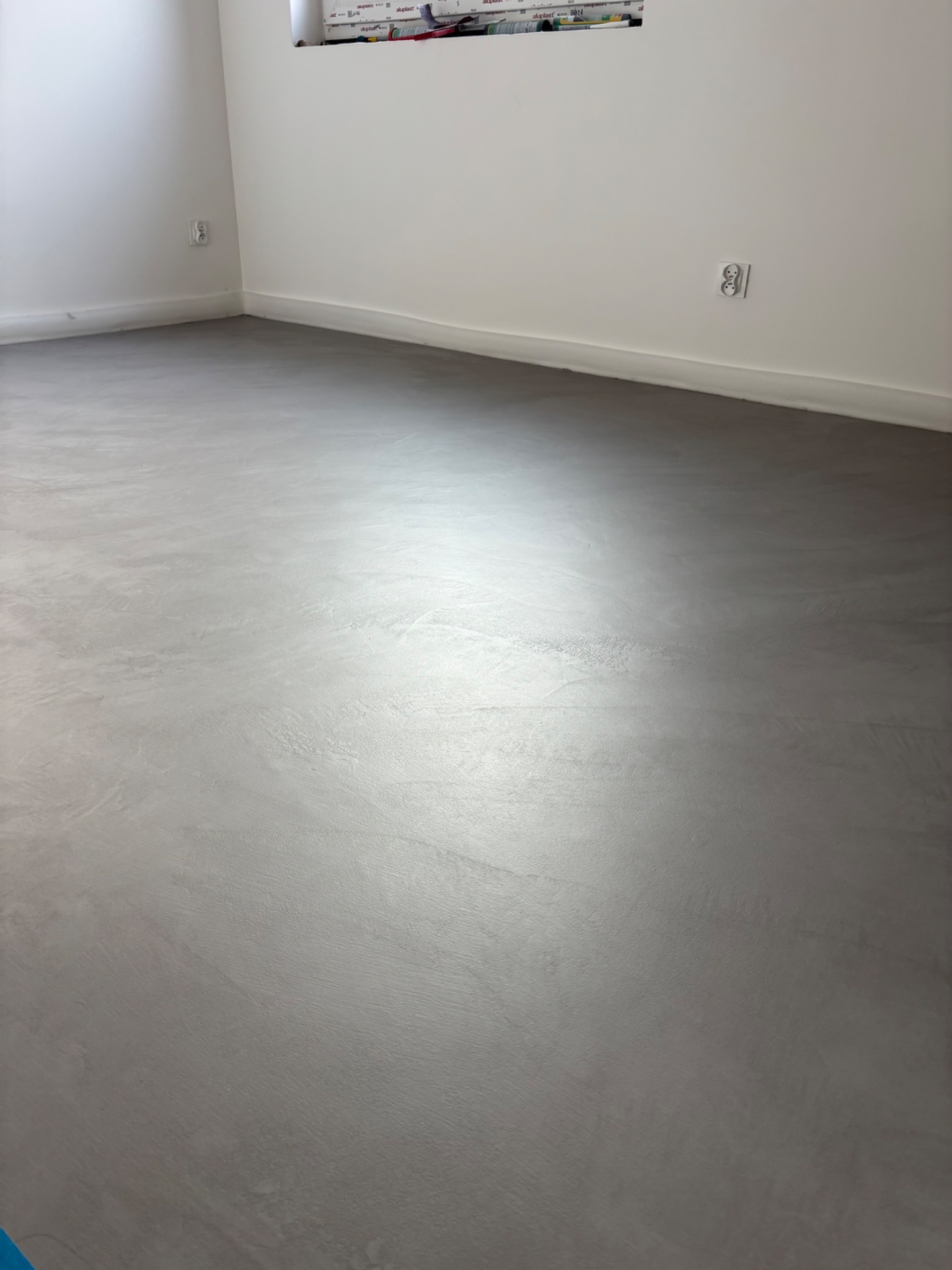 Szara, gładka posadzka w minimalistycznym wnętrzu. Widoczne gniazdka elektryczne na białej ścianie i listwa przypodłogowa. Delikatne refleksy światła na powierzchni podłogi.