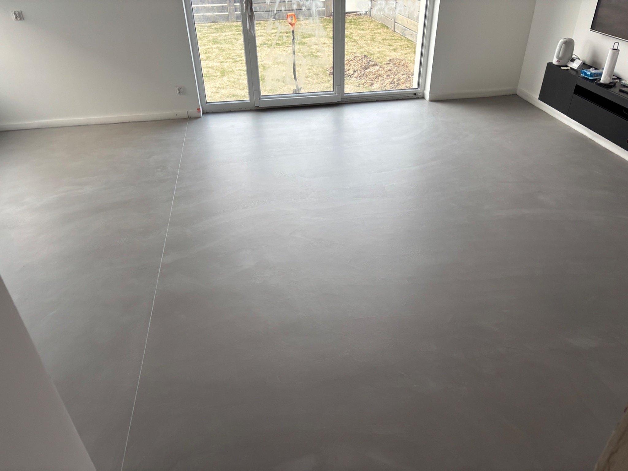 Szara, gładka posadzka w jasnym wnętrzu z dużym oknem na ogród. Minimalistyczny design, widoczna faktura materiału i delikatne refleksy światła.