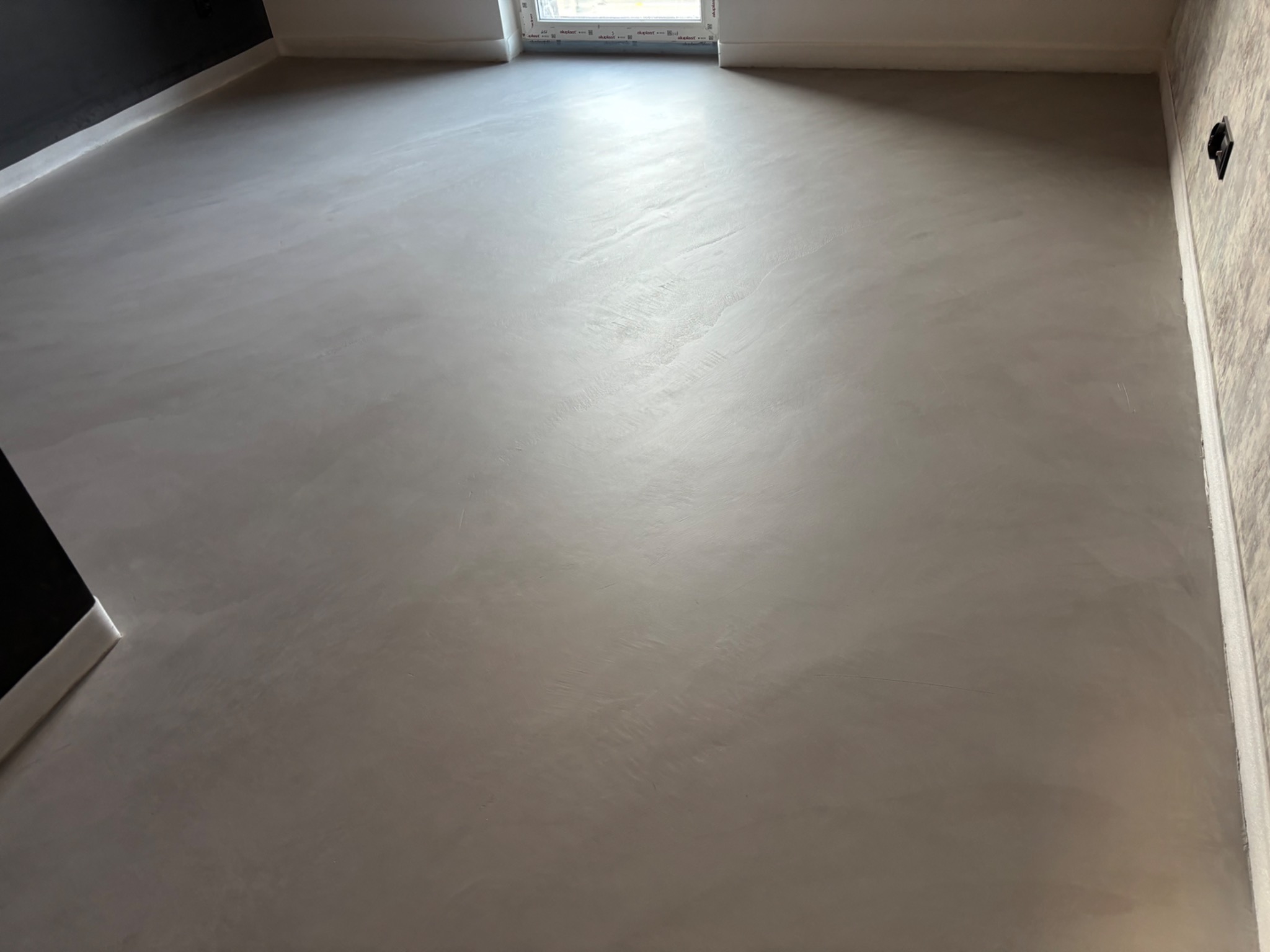 Jasnoszara, gładka posadzka w pokoju z oknem. Widoczne delikatne wzory i refleksy światła na powierzchni. Minimalistyczne wykończenie wnętrza.