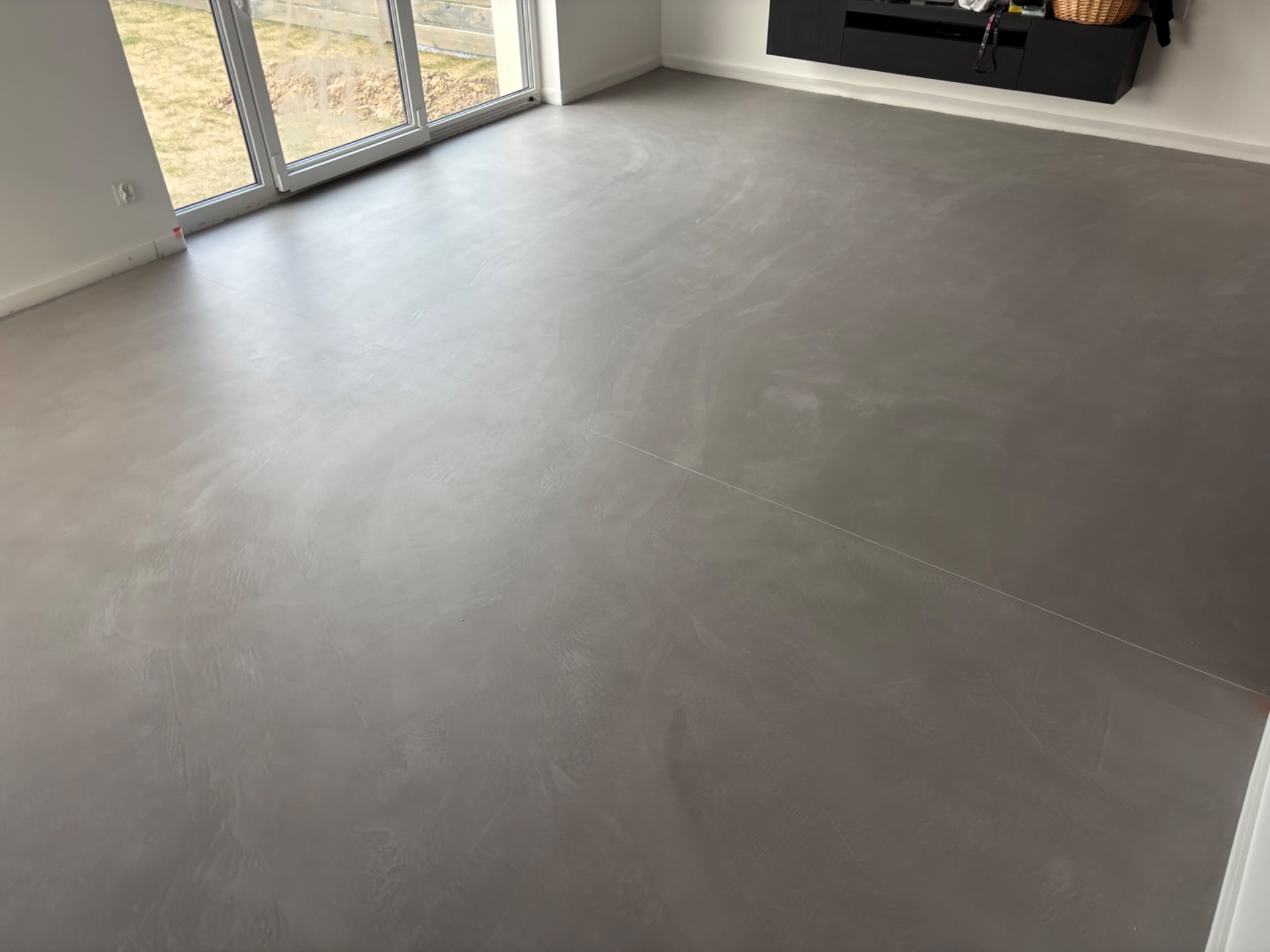 Szara, gładka posadzka z mikrocementu w jasnym pomieszczeniu z dużym oknem przesuwnym. Widoczne subtelne wzory i tekstura materiału. Minimalistyczny design wnętrza.