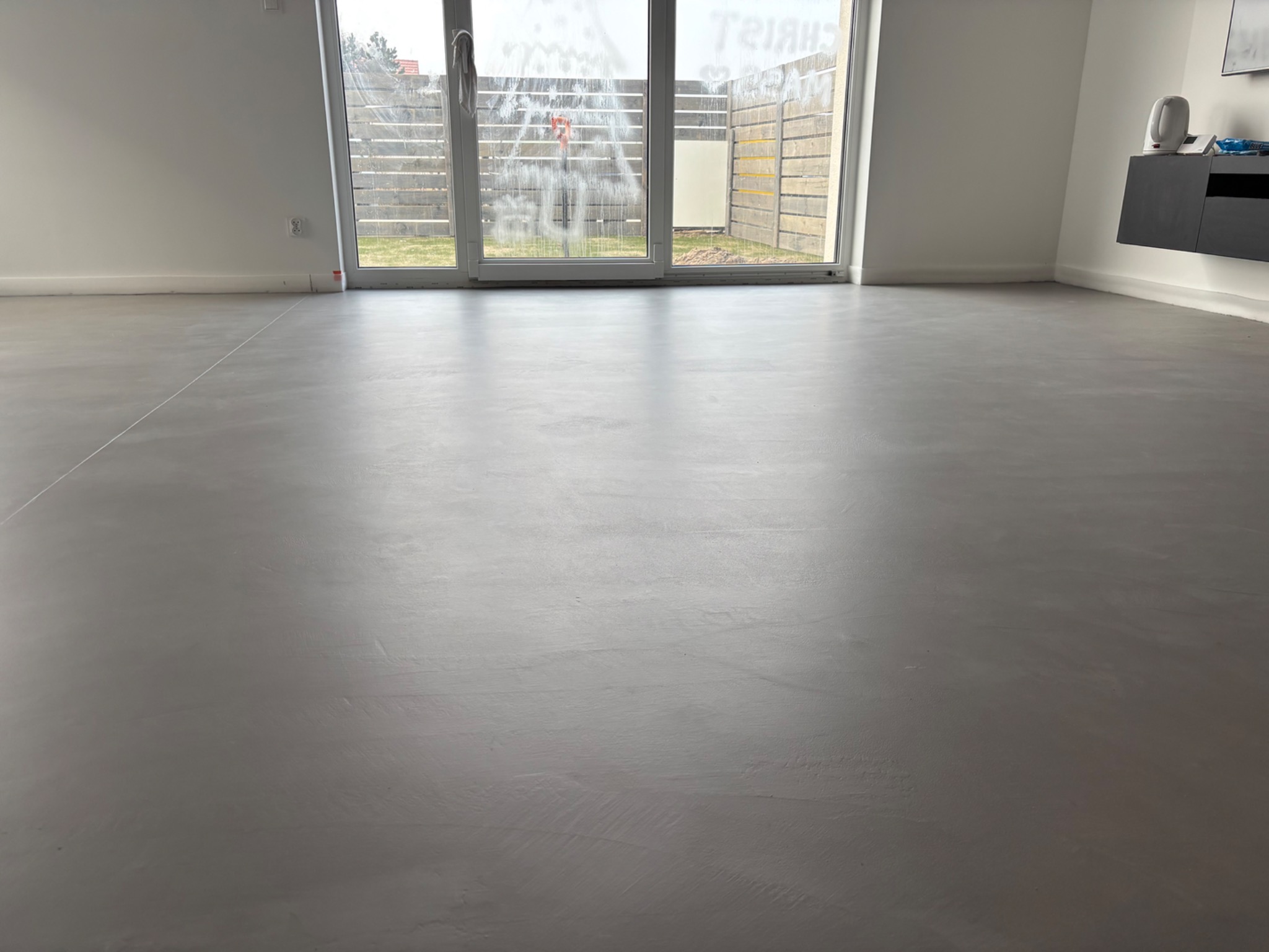 Szara, gładka podłoga w minimalistycznym wnętrzu z dużym oknem balkonowym i wiszącą szafką RTV. Widoczne delikatne smugi na powierzchni podłogi.
