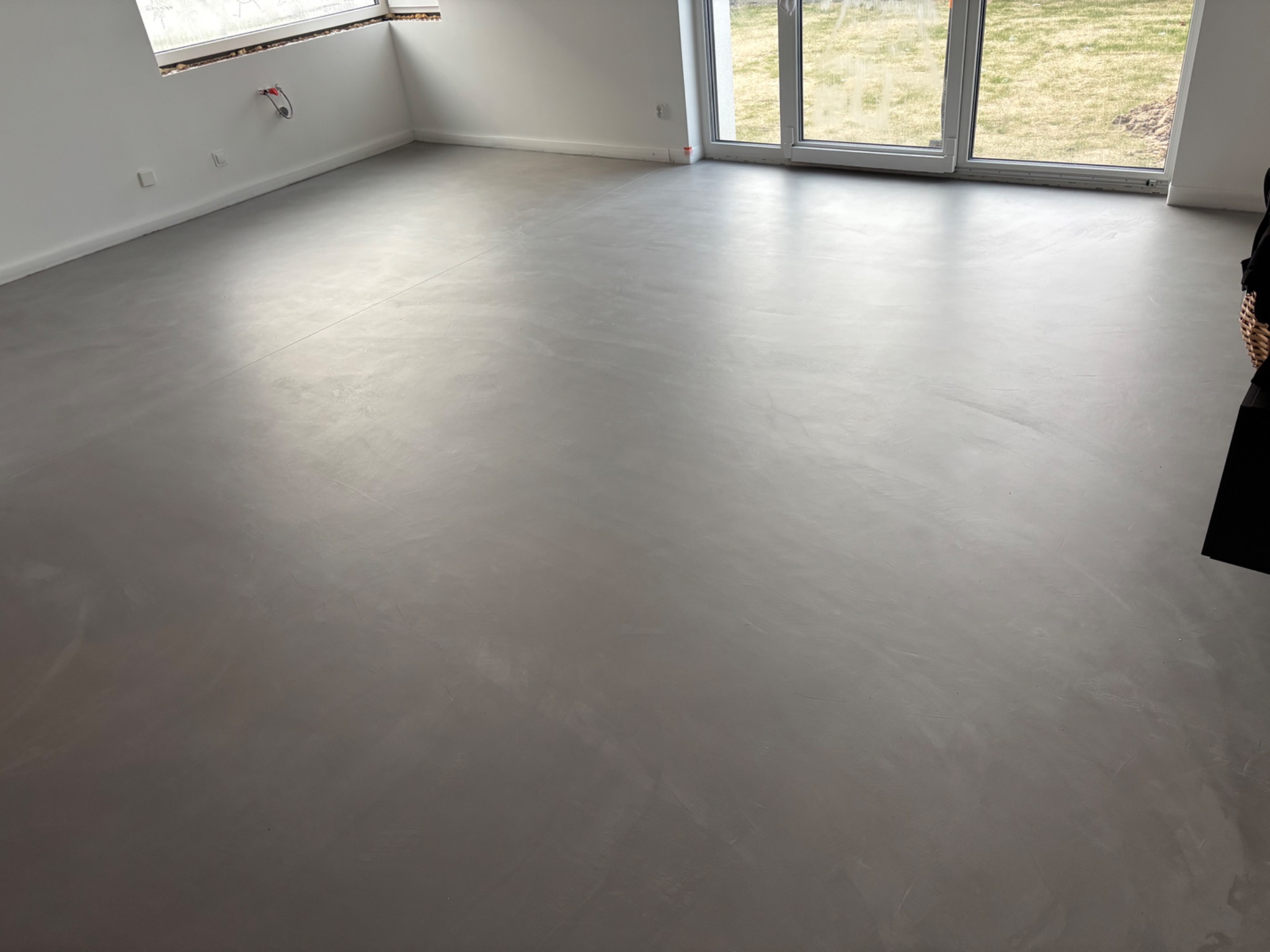 Szara, gładka posadzka w jasnym pomieszczeniu z dużymi oknami. Minimalistyczny design, widoczne subtelne wzory na powierzchni podłogi. Nowoczesne wykończenie wnętrza.