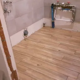 Ułożona podłoga z płytek imitujących drewno w trakcie prac wykończeniowych w łazience, widoczne instalacje wodne i fragmenty ścian w stanie surowym.