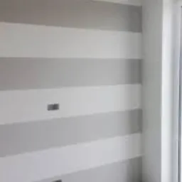 Ściana pomalowana w poziome pasy w odcieniach szarości i bieli, widoczne gniazdka elektryczne, kadr z fragmentem okna.