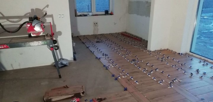 Układanie płytek podłogowych z użyciem klipsów poziomujących w jasnym pomieszczeniu, widoczna przecinarka do płytek.