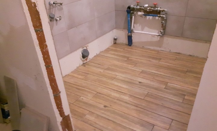 Ułożona podłoga z płytek imitujących drewno w trakcie prac wykończeniowych w łazience, widoczne instalacje wodne i fragmenty ścian w stanie surowym.